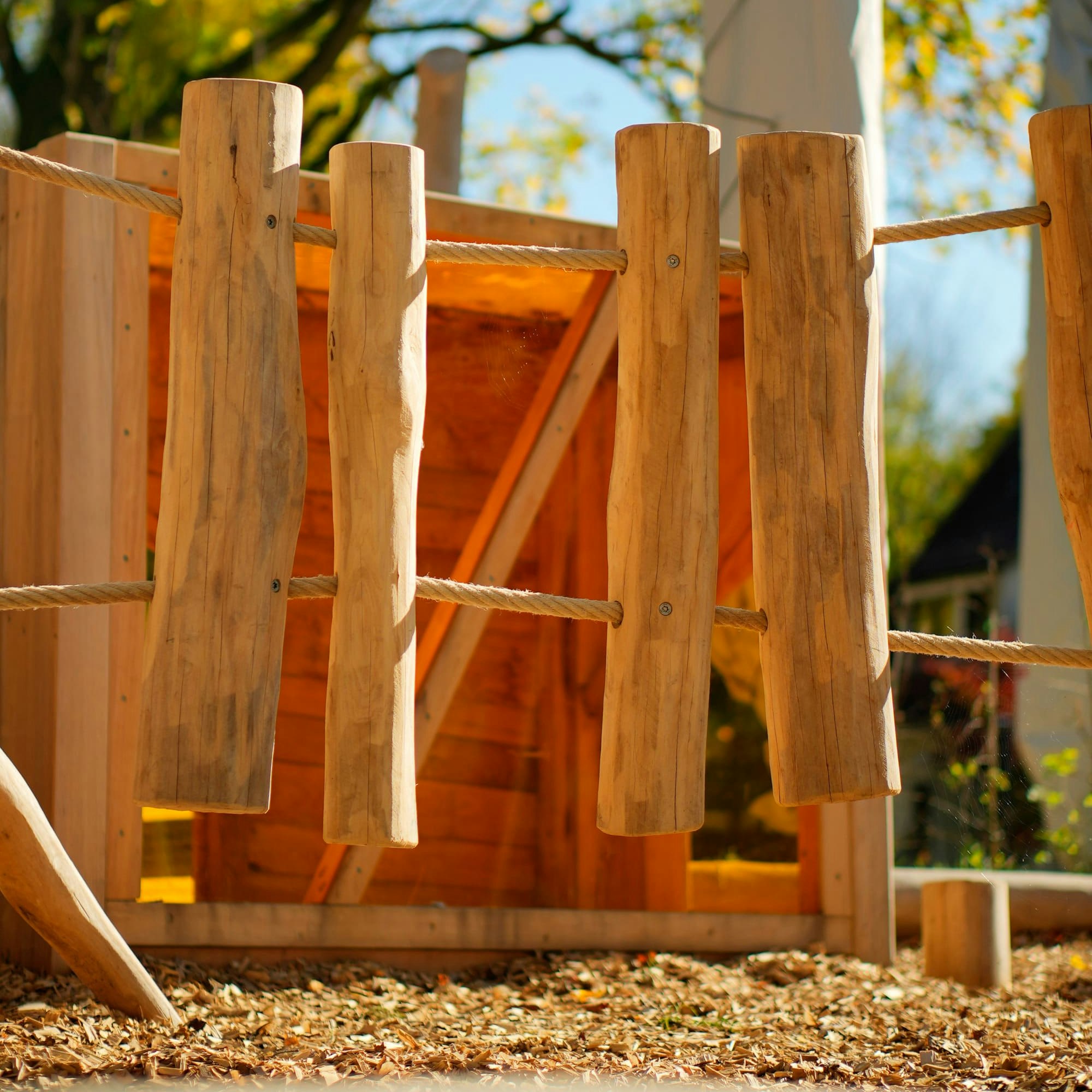 Ein hölzernes Spielgerüst für Kinder mit senkrechten Baumstämmen und einem Seil, umgeben von Mulch, vor einem Hintergrund aus Bäumen mit Herbstlaub.