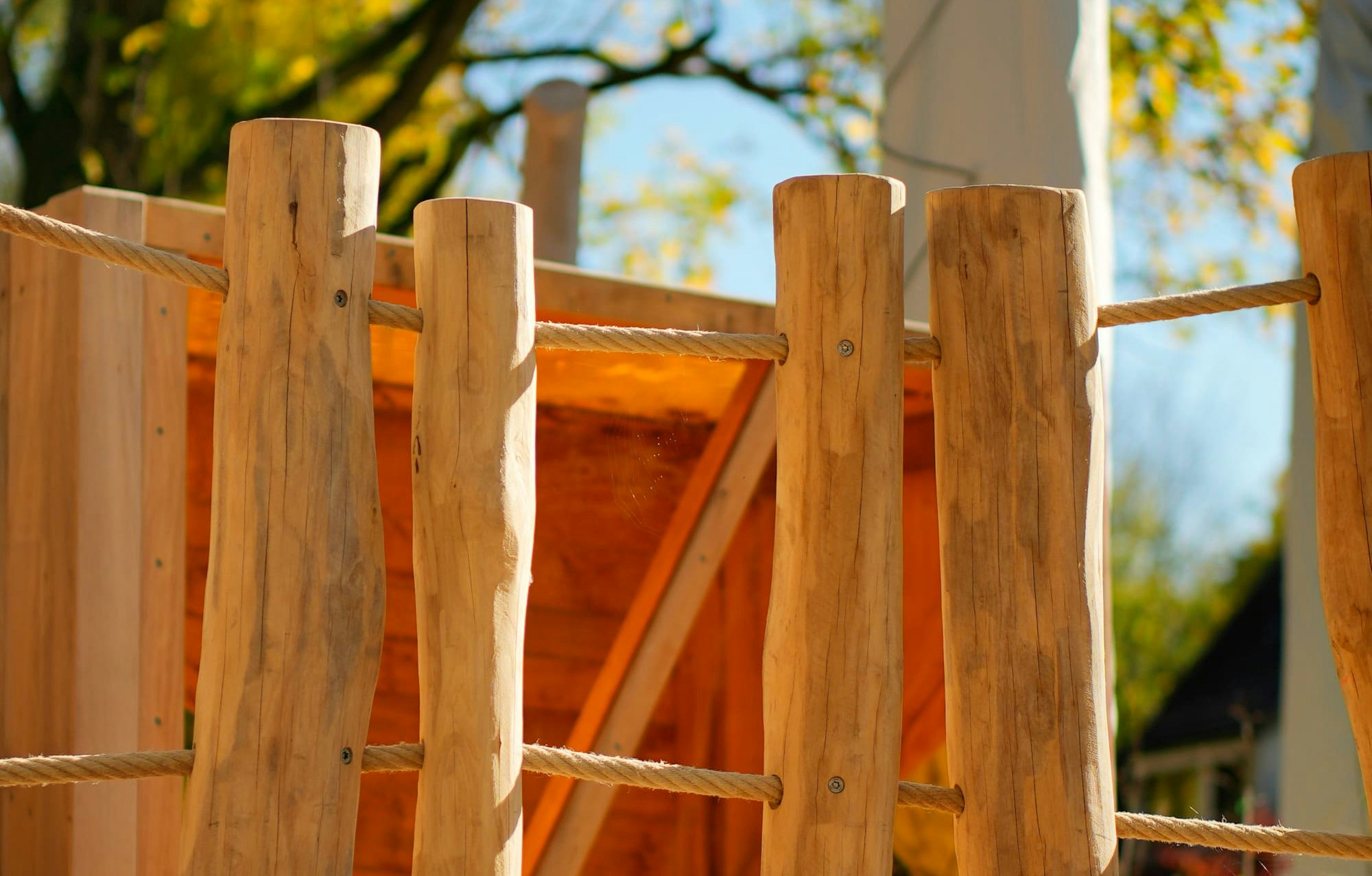 Ein hölzernes Spielgerüst für Kinder mit senkrechten Baumstämmen und einem Seil, umgeben von Mulch, vor einem Hintergrund aus Bäumen mit Herbstlaub.