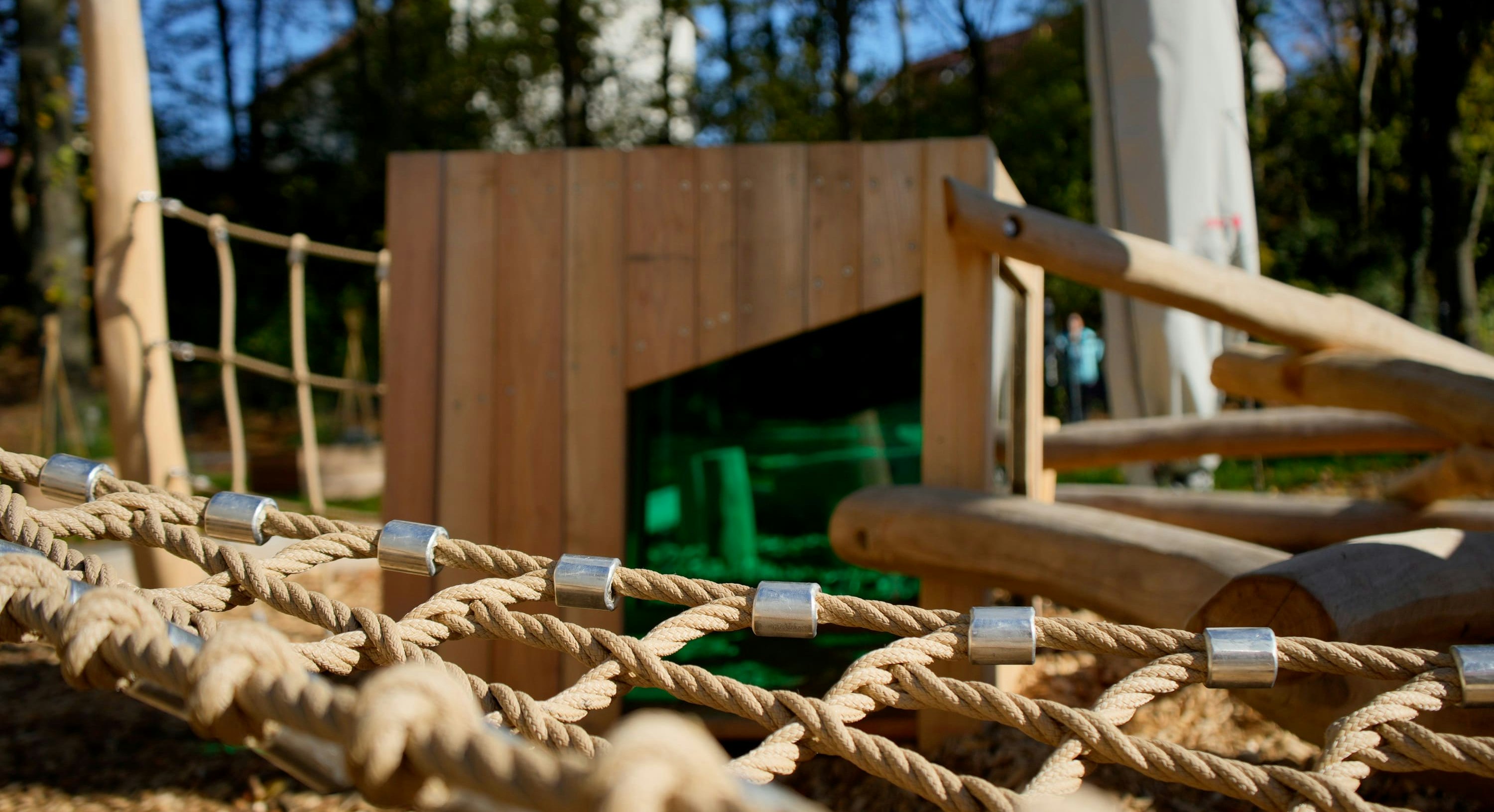 Nahaufnahme einer Seilnetz-Spielstruktur auf einem Spielplatz mit einer hölzernen Kletterwand und einer grünen Rutsche im Hintergrund. Dahinter sind Bäume und ein weißes Gebäude zu sehen.