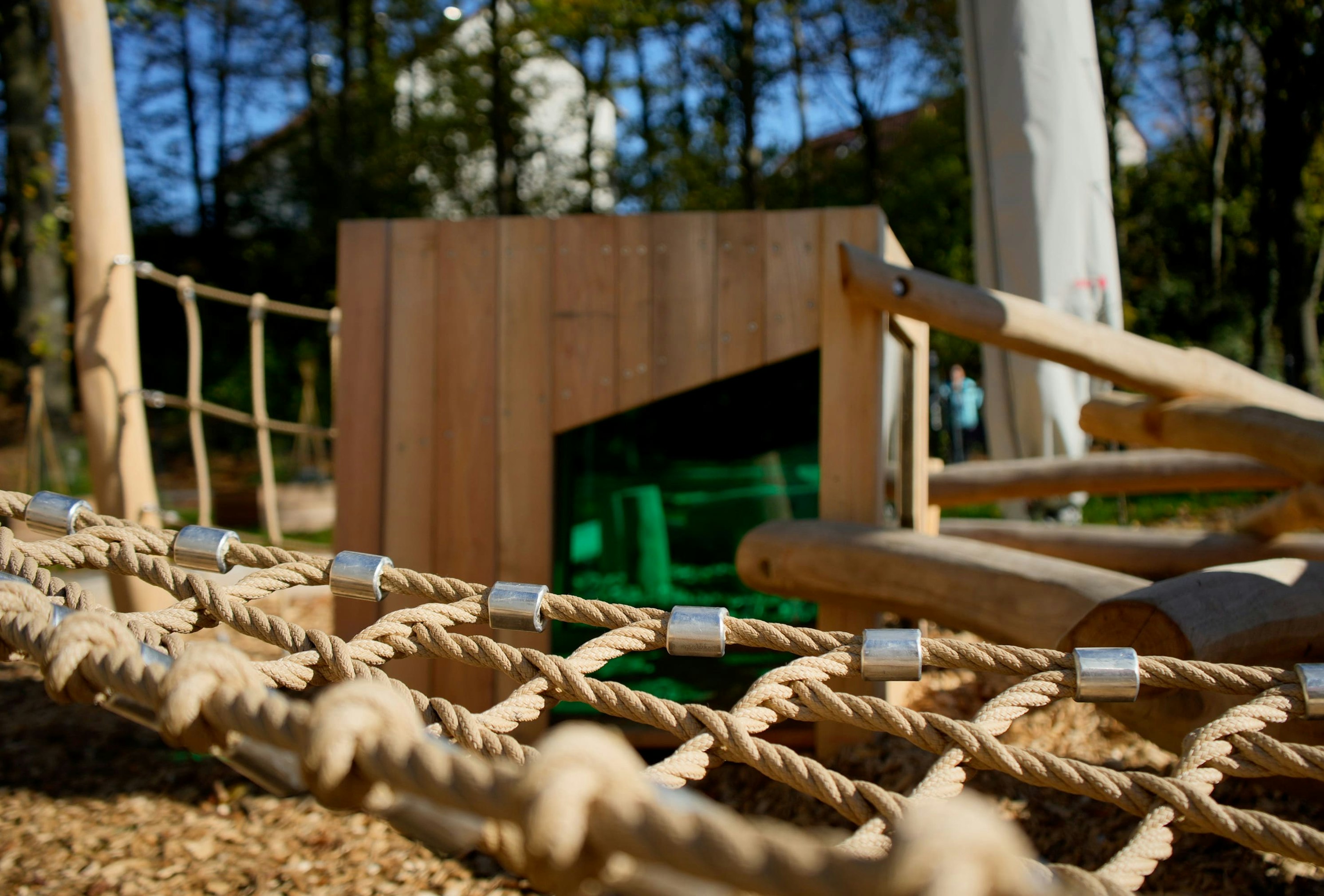 Nahaufnahme einer Seilnetz-Spielstruktur auf einem Spielplatz mit einer hölzernen Kletterwand und einer grünen Rutsche im Hintergrund. Dahinter sind Bäume und ein weißes Gebäude zu sehen.