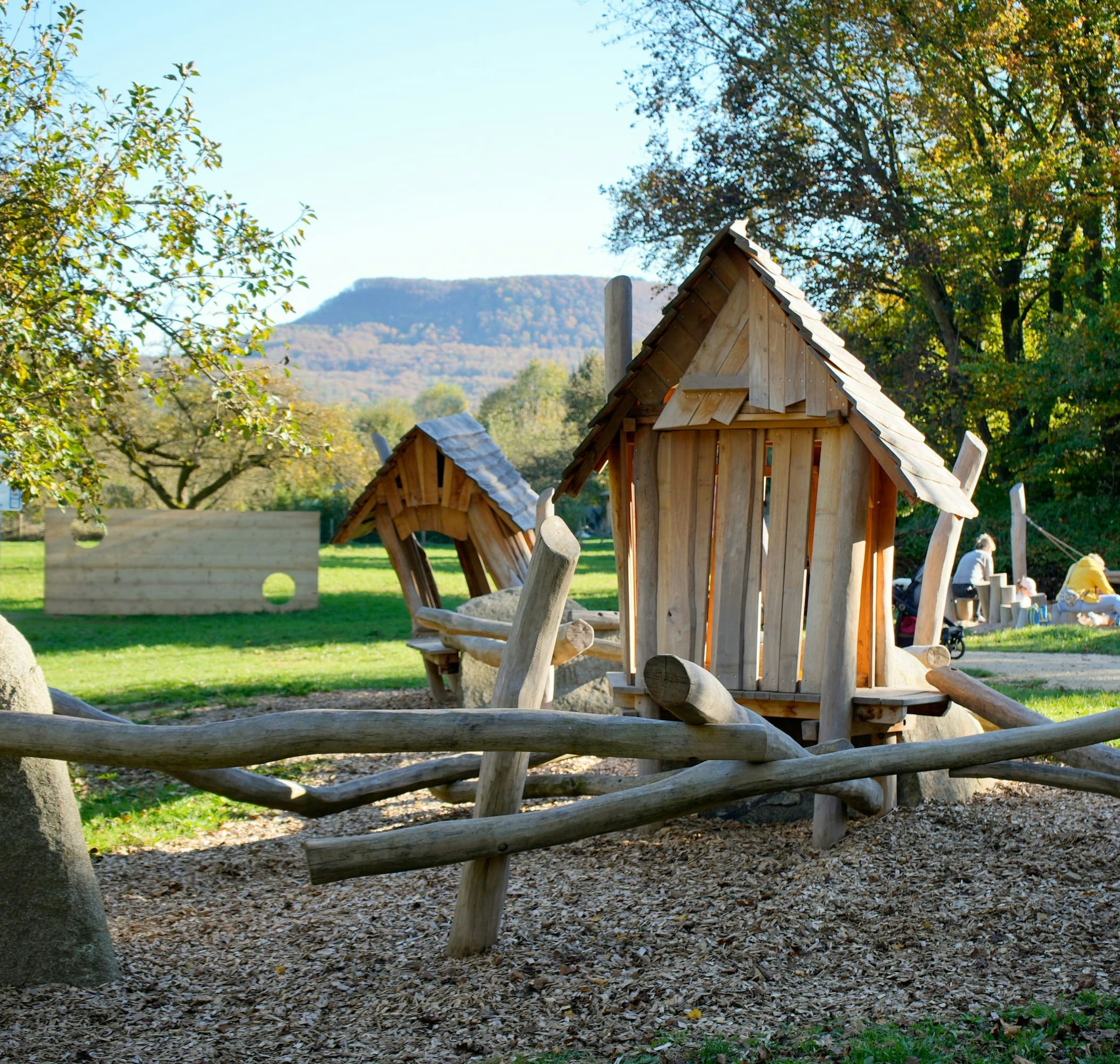 Eine hölzerne Spielplatzstruktur mit Hütten und Baumstämmen ist von Bäumen und Gras umgeben. Im Hintergrund sind Menschen zu sehen.