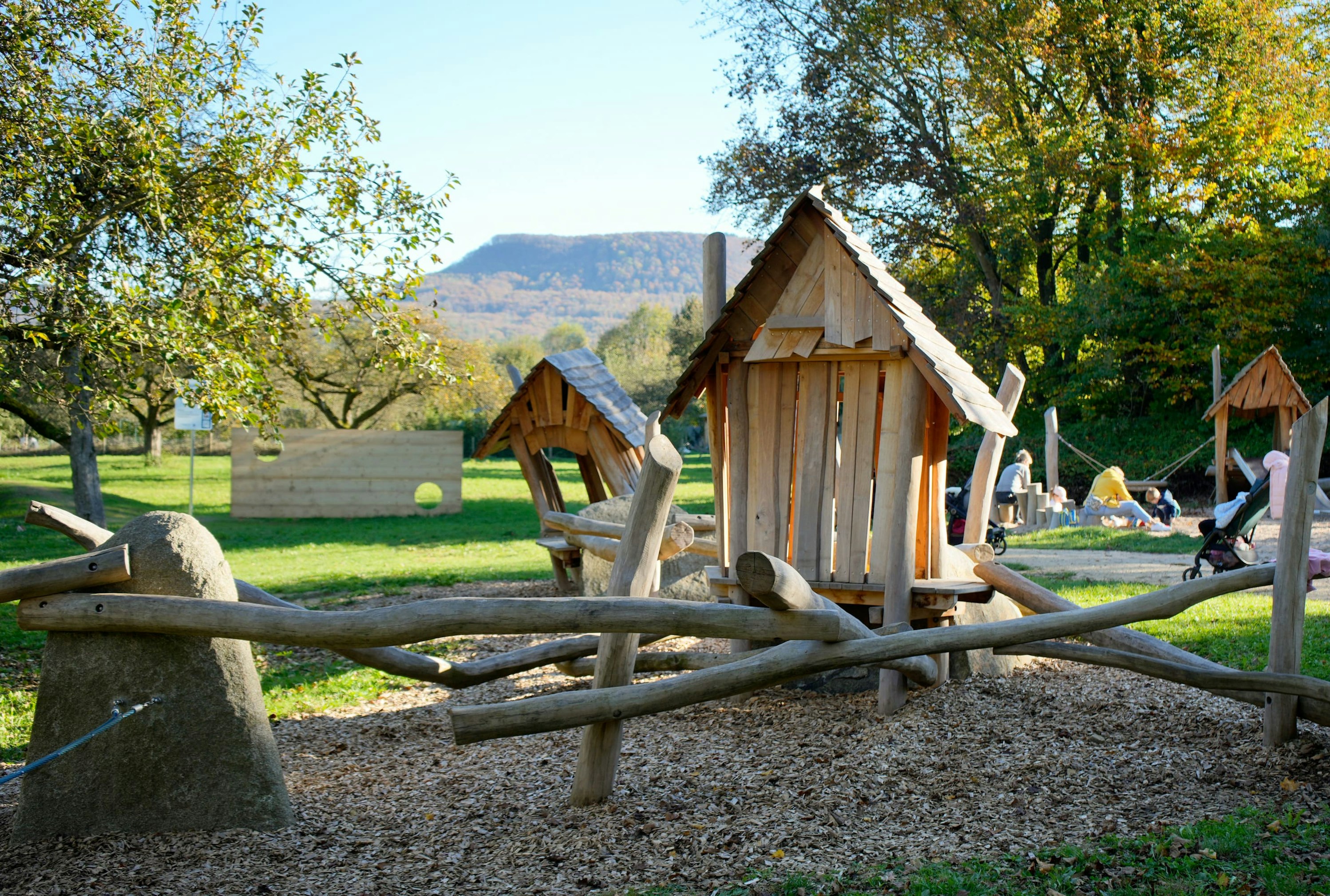 Eine hölzerne Spielplatzstruktur mit Hütten und Baumstämmen ist von Bäumen und Gras umgeben. Im Hintergrund sind Menschen zu sehen.