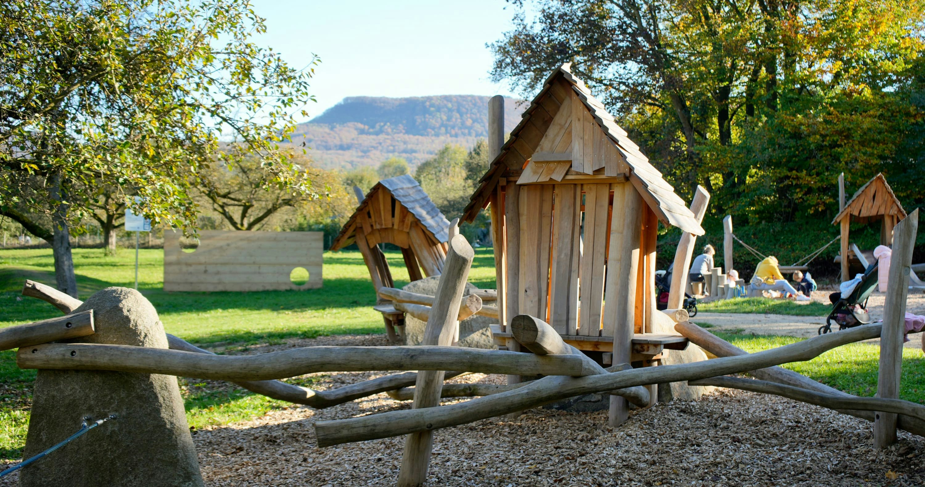 Eine hölzerne Spielplatzstruktur mit Hütten und Baumstämmen ist von Bäumen und Gras umgeben. Im Hintergrund sind Menschen zu sehen.