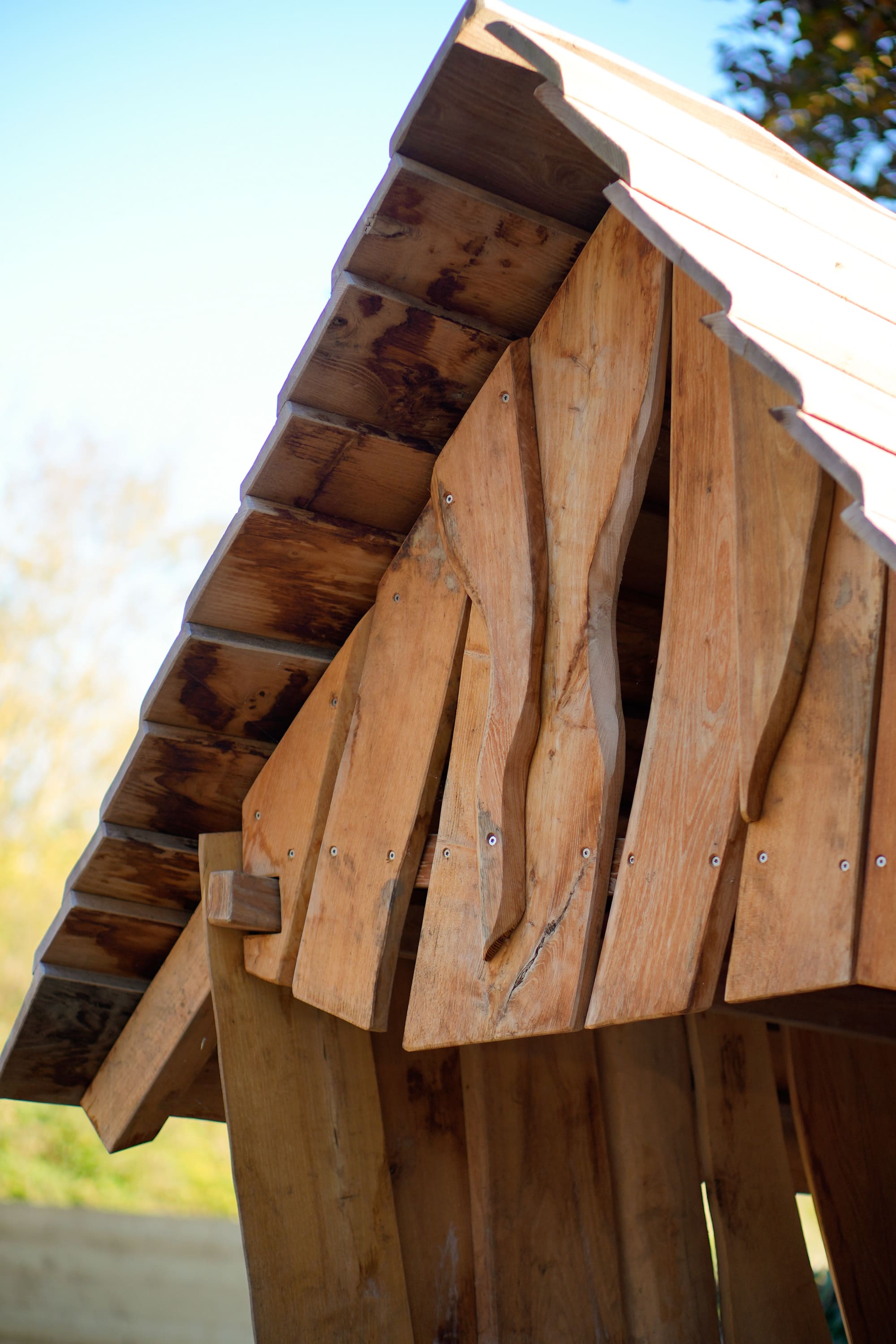 Nahaufnahme einer Holzkonstruktion mit überlappenden, unregelmäßig geformten Latten und einem Satteldach vor einem blauen Himmel.