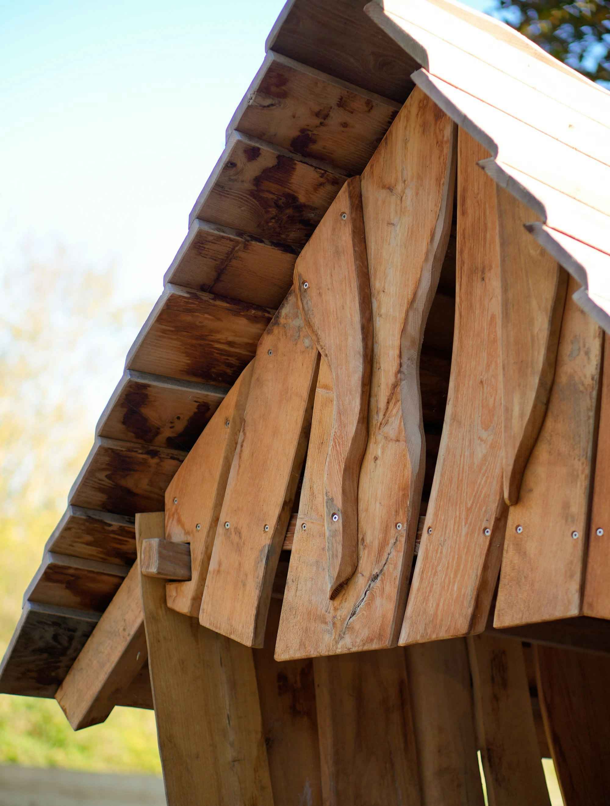 Nahaufnahme einer Holzkonstruktion mit überlappenden, unregelmäßig geformten Latten und einem Satteldach vor einem blauen Himmel.