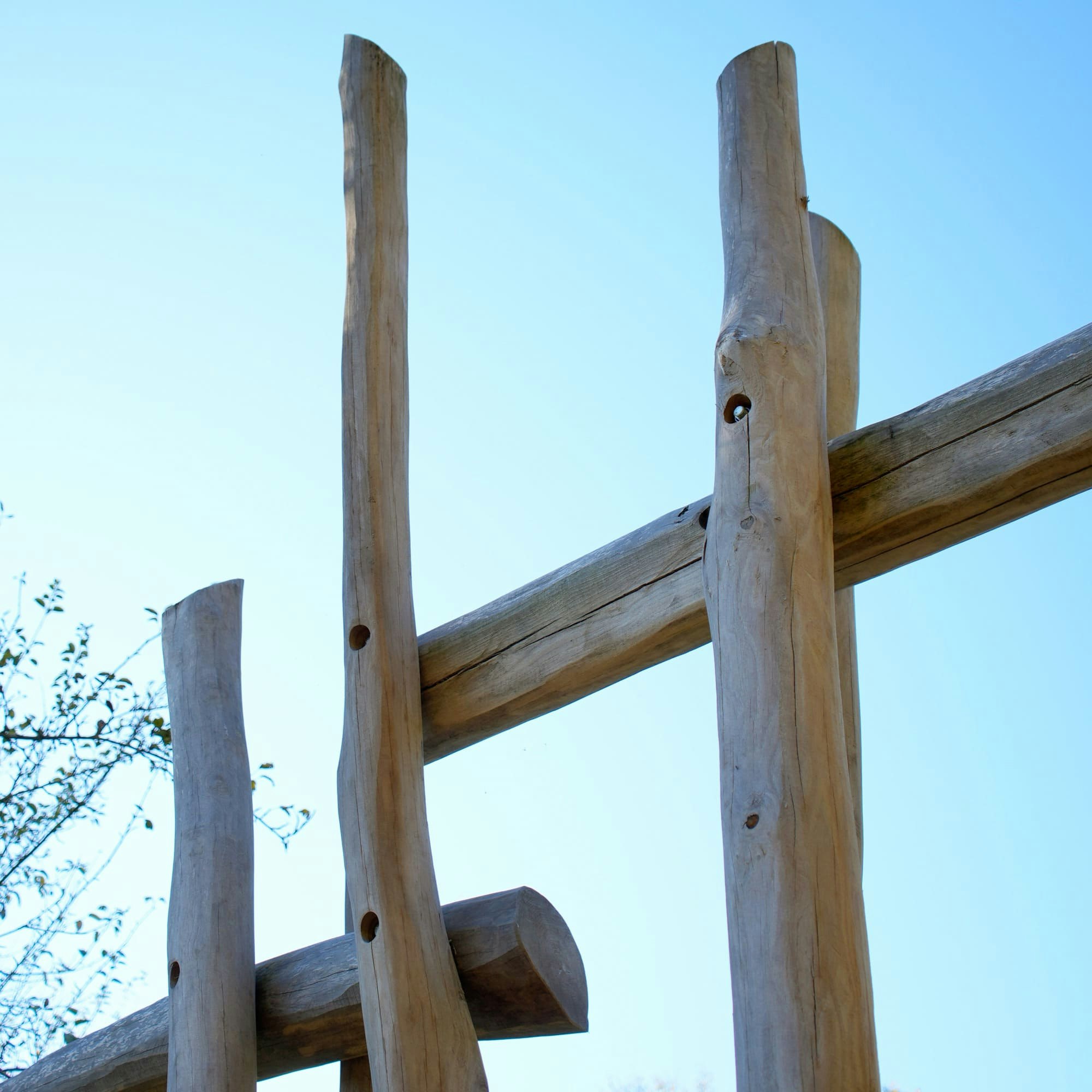 Hohe, vertikal und horizontal angeordnete Holzbalken vor einem klaren blauen Himmel mit Bäumen im Hintergrund.