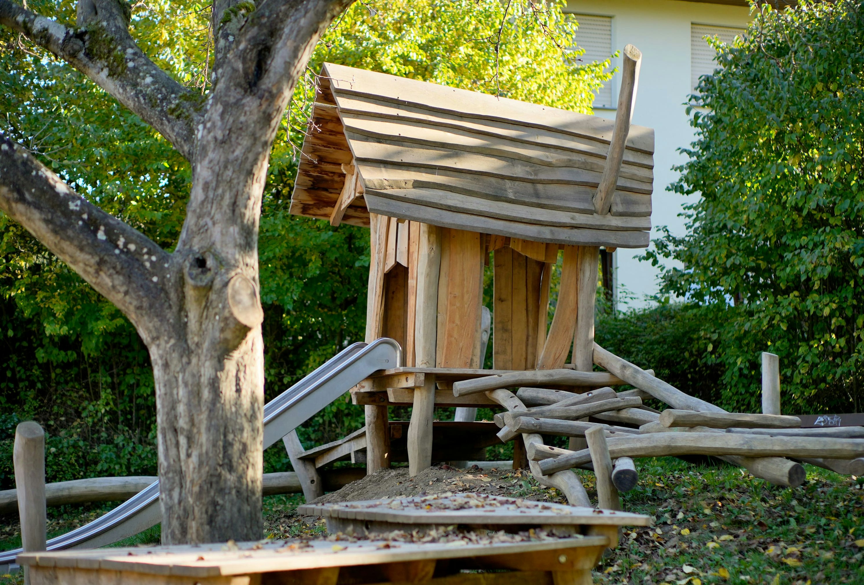 Hölzerne Spielplatzstruktur mit Rutsche und Kletterelementen unter einem Baum, umgeben von Herbstlaub.