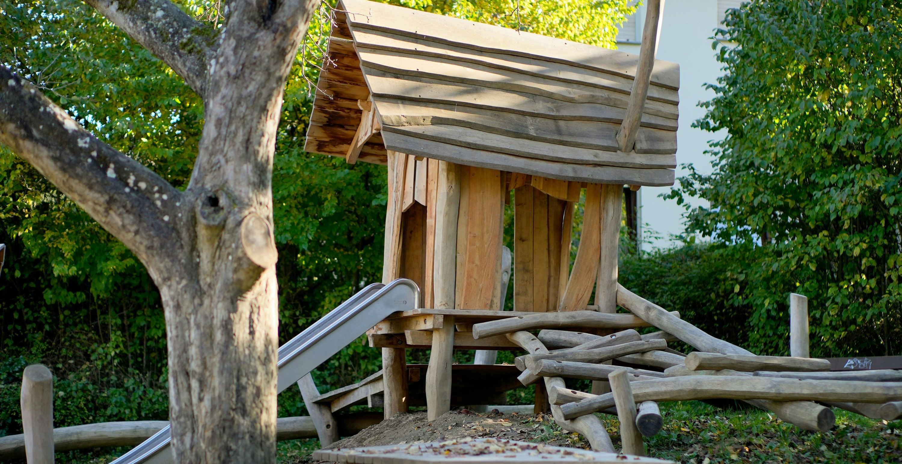 Hölzerne Spielplatzstruktur mit Rutsche und Kletterelementen unter einem Baum, umgeben von Herbstlaub.