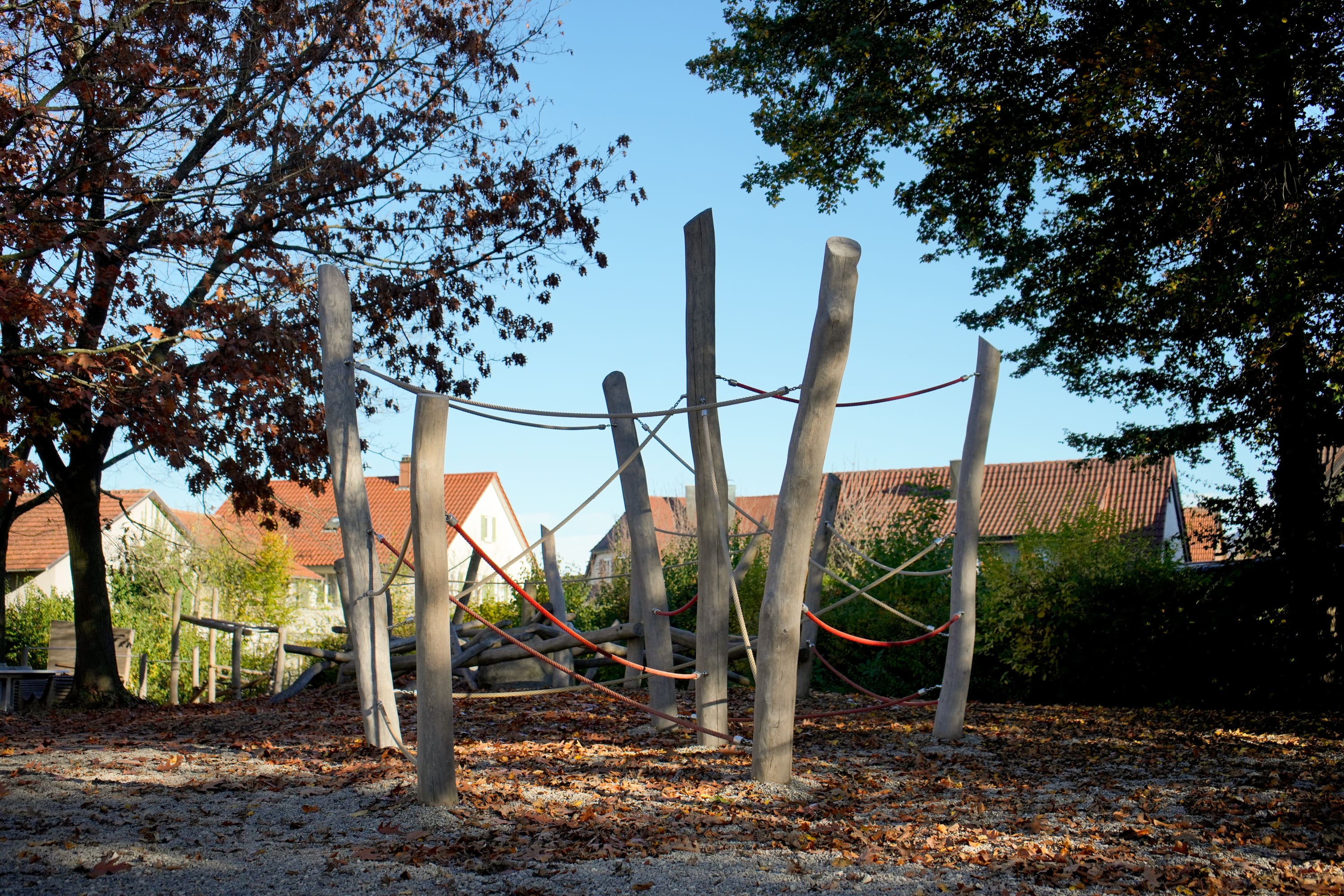 Außenspielplatz mit hohen Holzstangen und Seilen zum Klettern, umgeben von Bäumen mit Herbstlaub und Häusern im Hintergrund.
