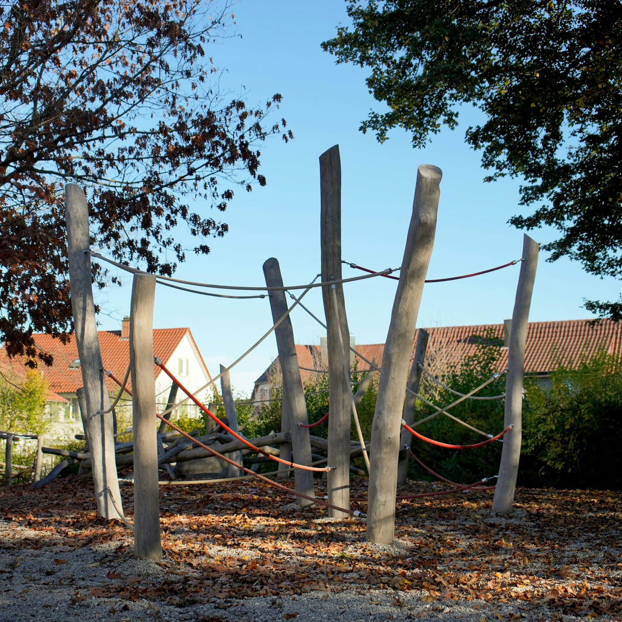 Außenspielplatz mit hohen Holzstangen und Seilen zum Klettern, umgeben von Bäumen mit Herbstlaub und Häusern im Hintergrund.