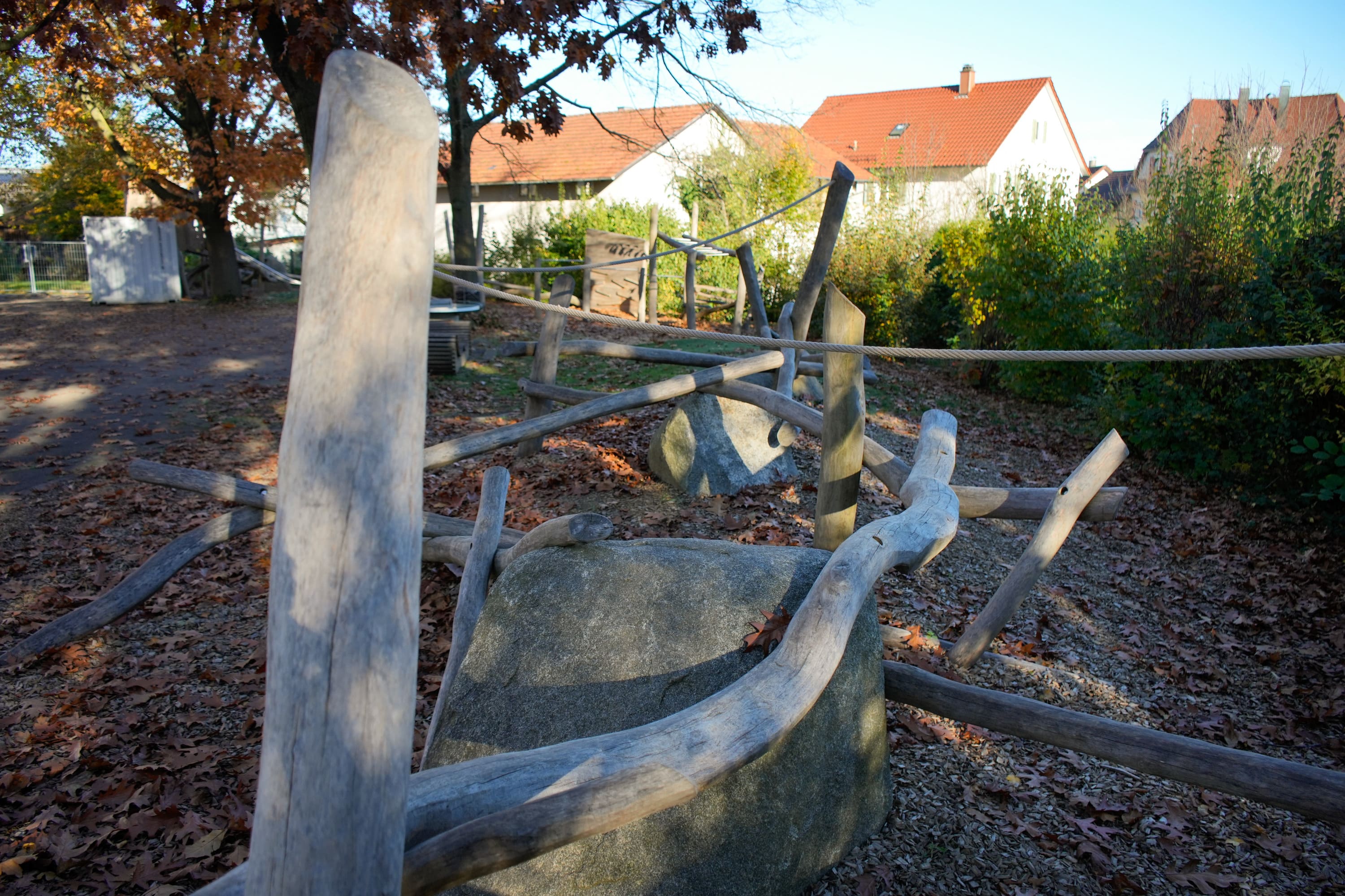 Hölzernes Klettergerüst auf einem Spielplatz mit Seilen und Baumstämmen, umgeben von Bäumen und nahe gelegenen Häusern.