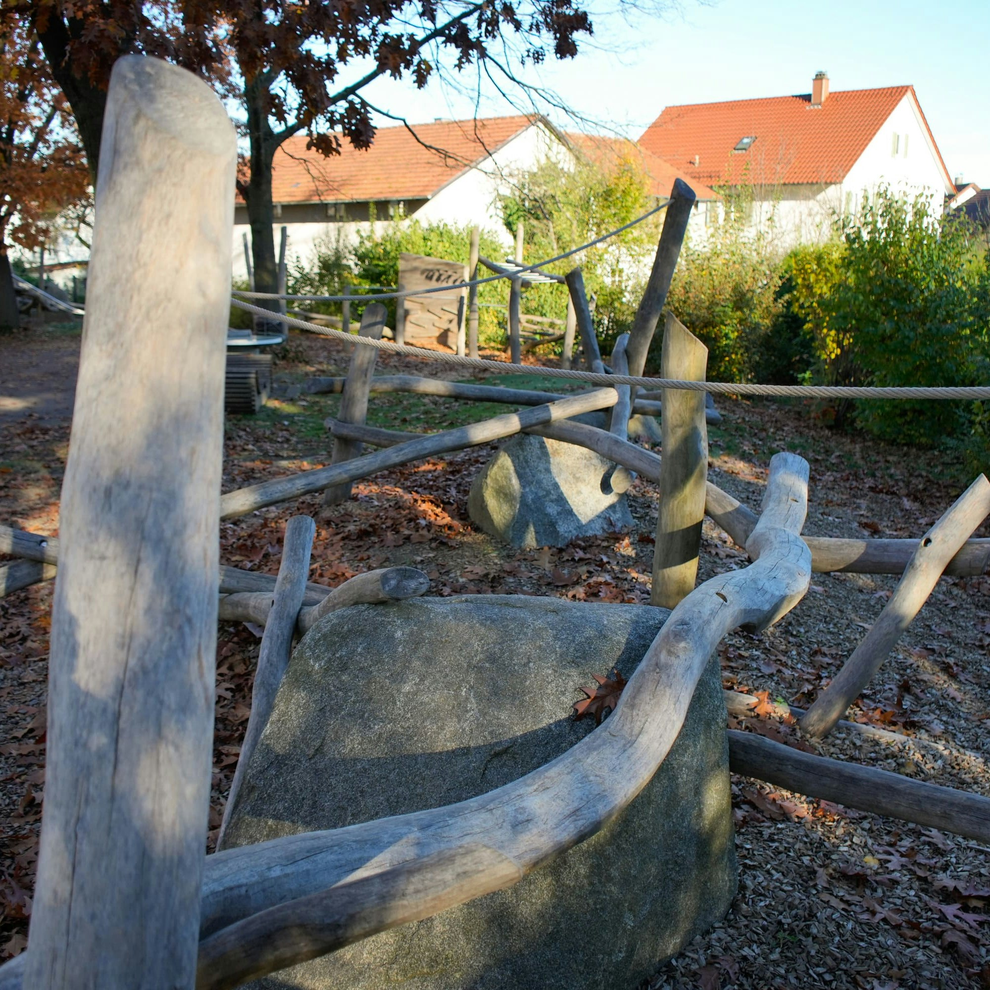 Hölzernes Klettergerüst auf einem Spielplatz mit Seilen und Baumstämmen, umgeben von Bäumen und nahe gelegenen Häusern.