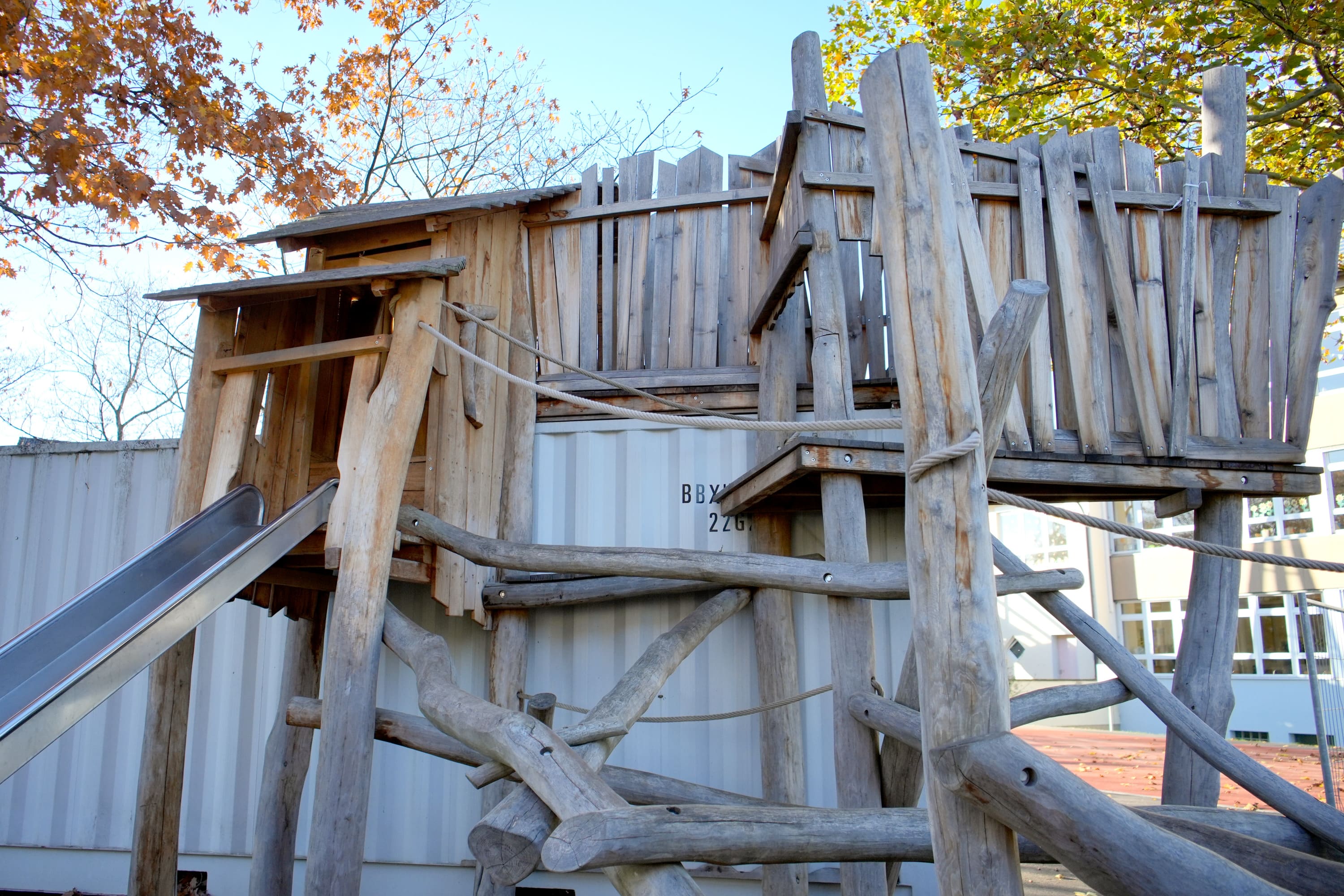 Holzspielgerät mit Rutsche und Plattformen aus grob behauenen Baumstämmen. Im Hintergrund sind weiße Container und herbstliche Bäume zu sehen.
