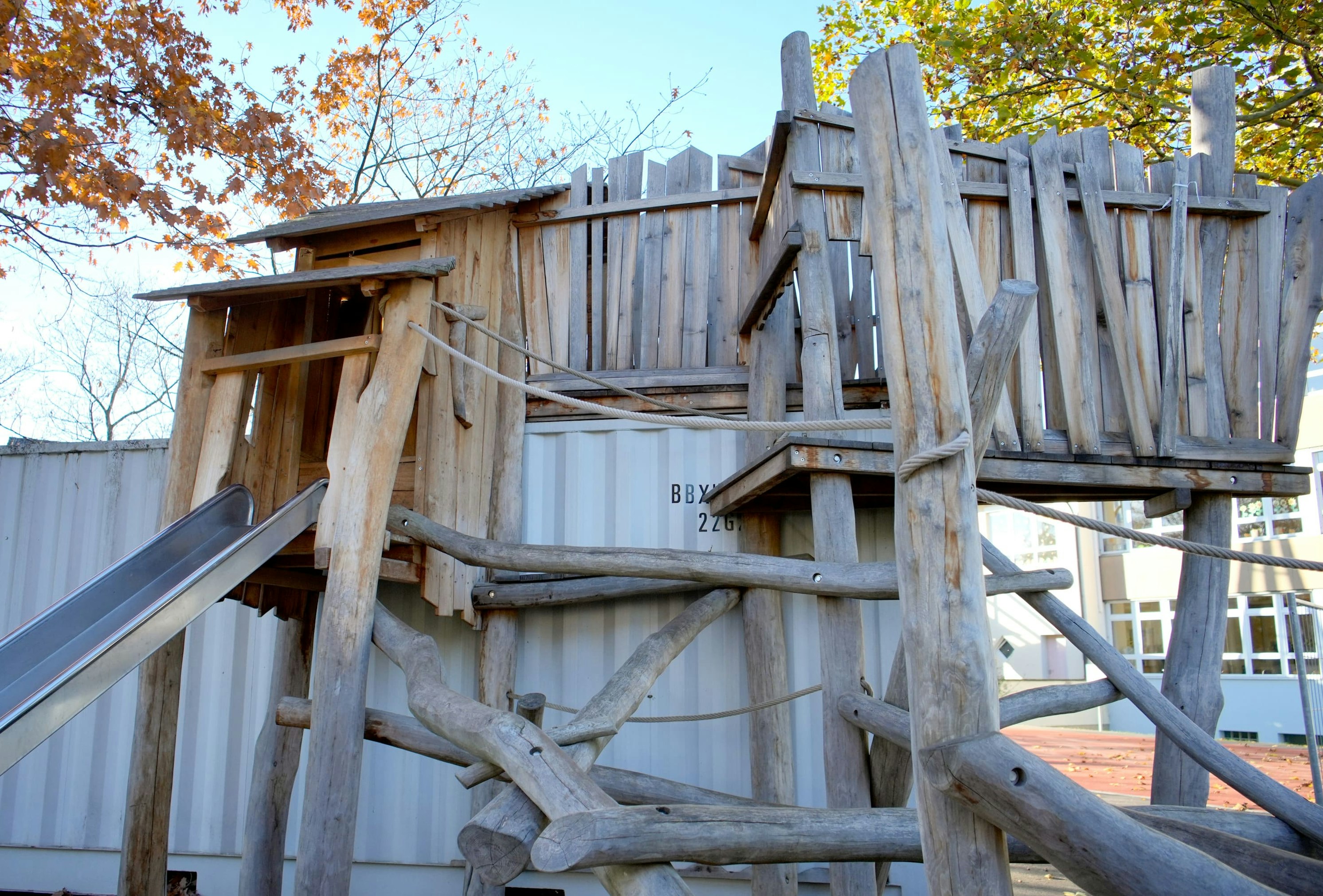 Holzspielgerät mit Rutsche und Plattformen aus grob behauenen Baumstämmen. Im Hintergrund sind weiße Container und herbstliche Bäume zu sehen.