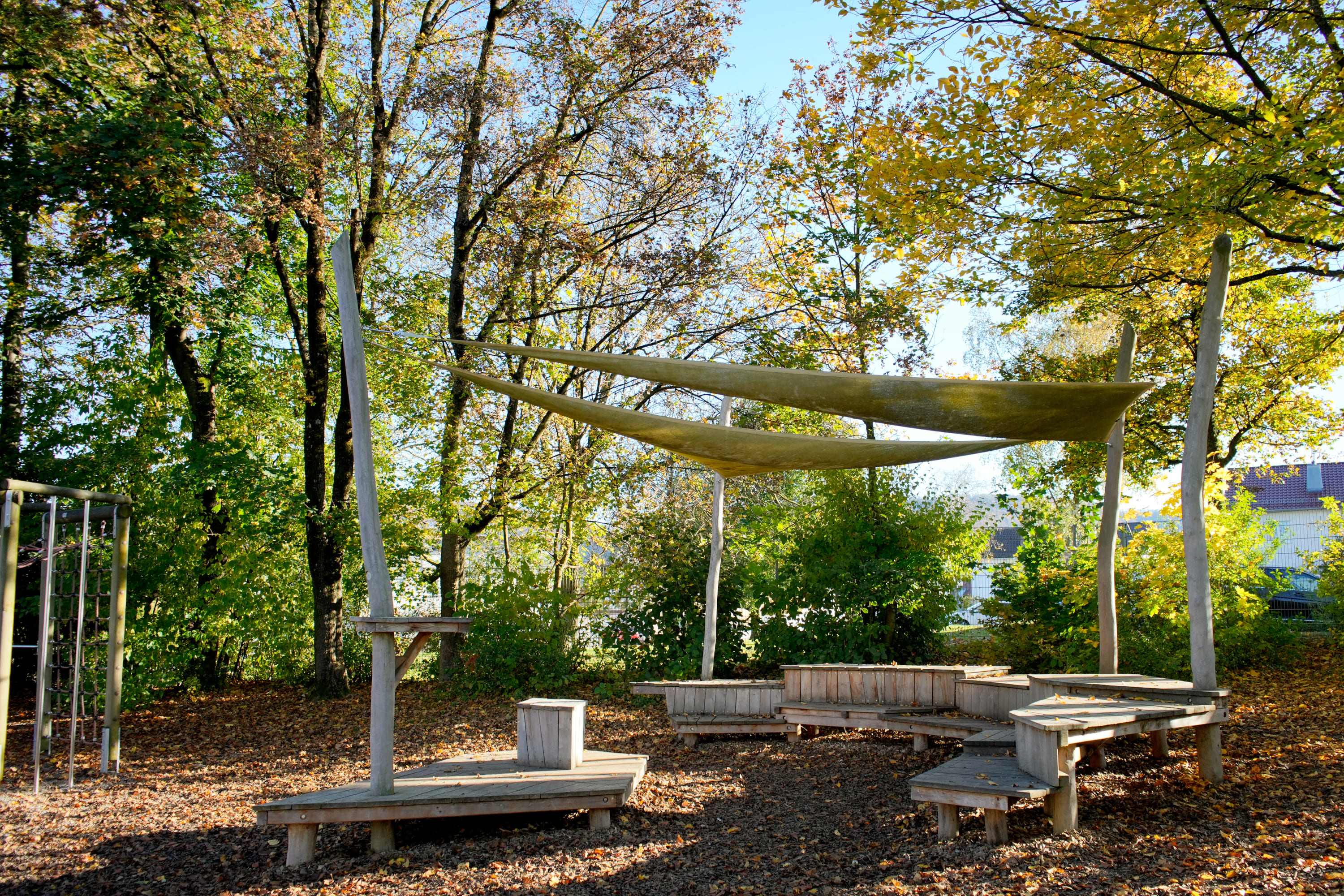 Außensitzbereich mit Holzbänken und Sonnensegel, umgeben von Bäumen mit grünen und gelben Blättern.