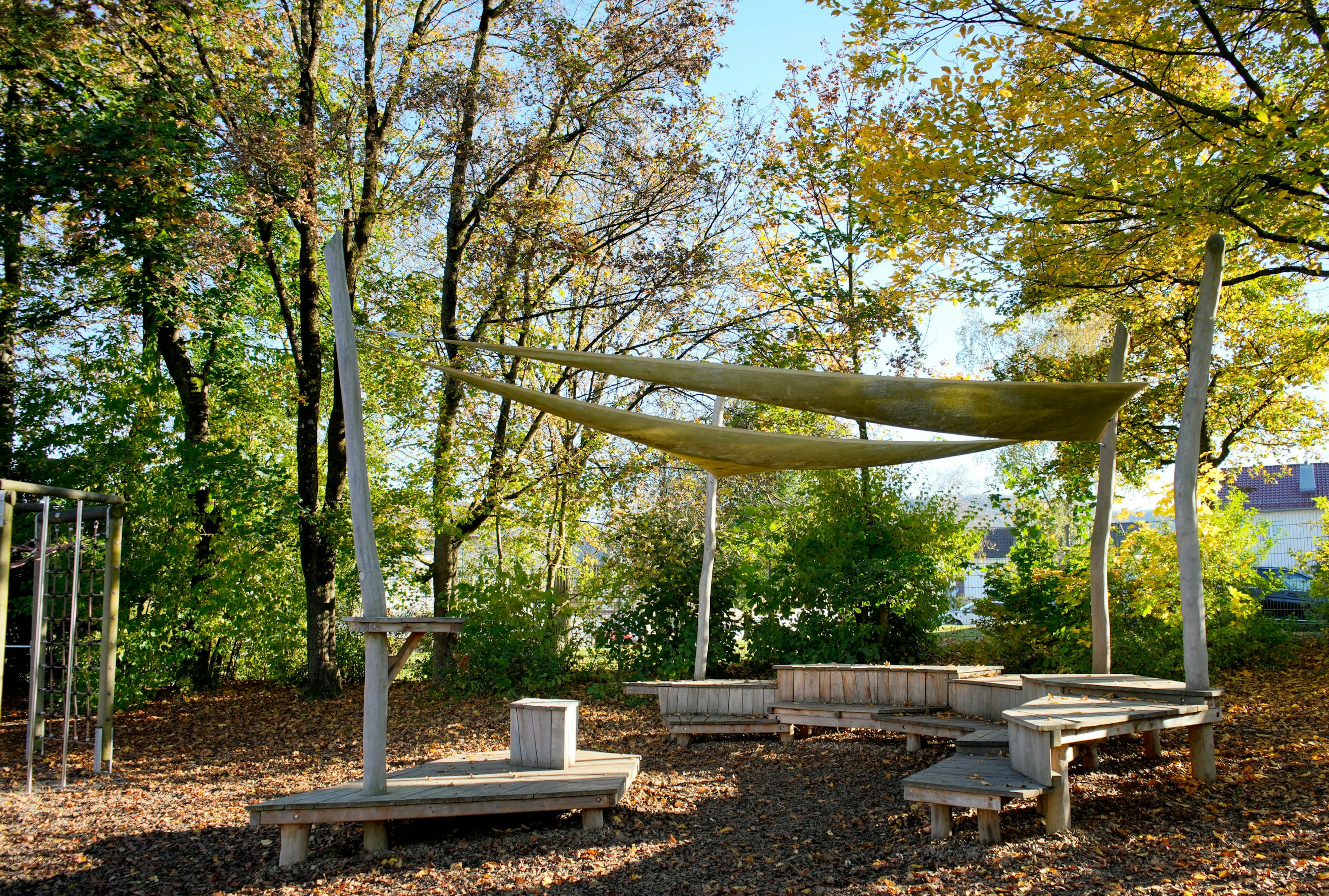 Außensitzbereich mit Holzbänken und Sonnensegel, umgeben von Bäumen mit grünen und gelben Blättern.