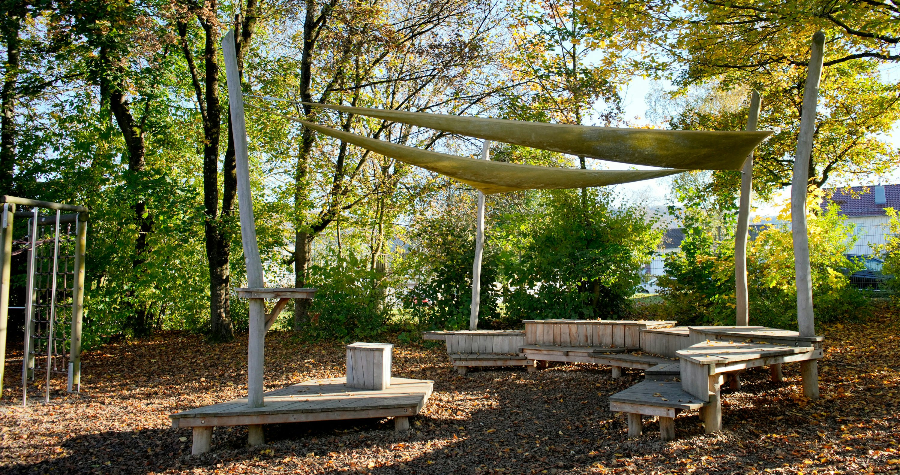 Außensitzbereich mit Holzbänken und Sonnensegel, umgeben von Bäumen mit grünen und gelben Blättern.