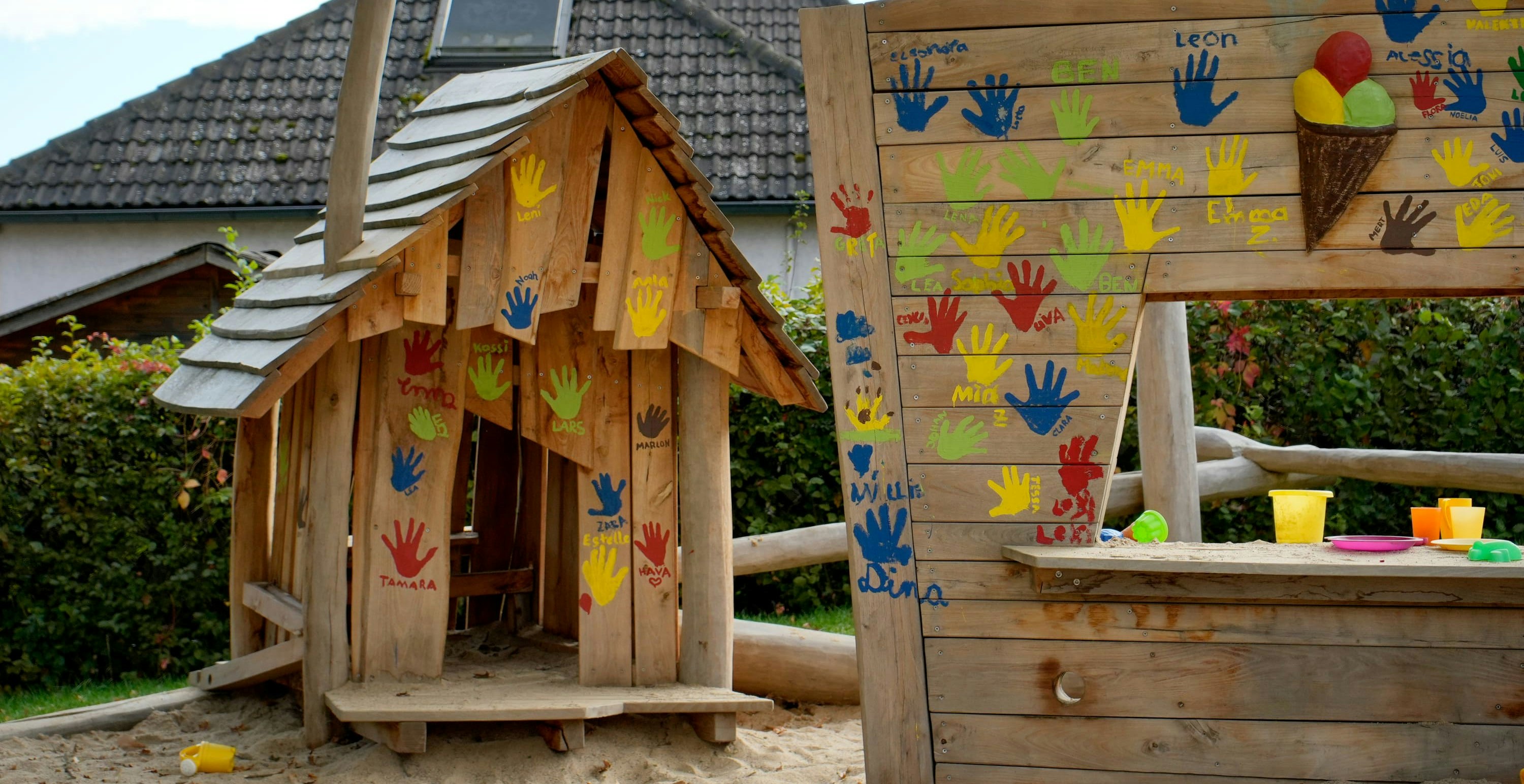 Ein hölzernes Spielhaus und ein Zaun mit bunten Handabdrücken und Namen. Im Hintergrund ein kleiner Hof, in dem ein Haus zu sehen ist. Plastikspielzeug liegt verstreut im Sand.