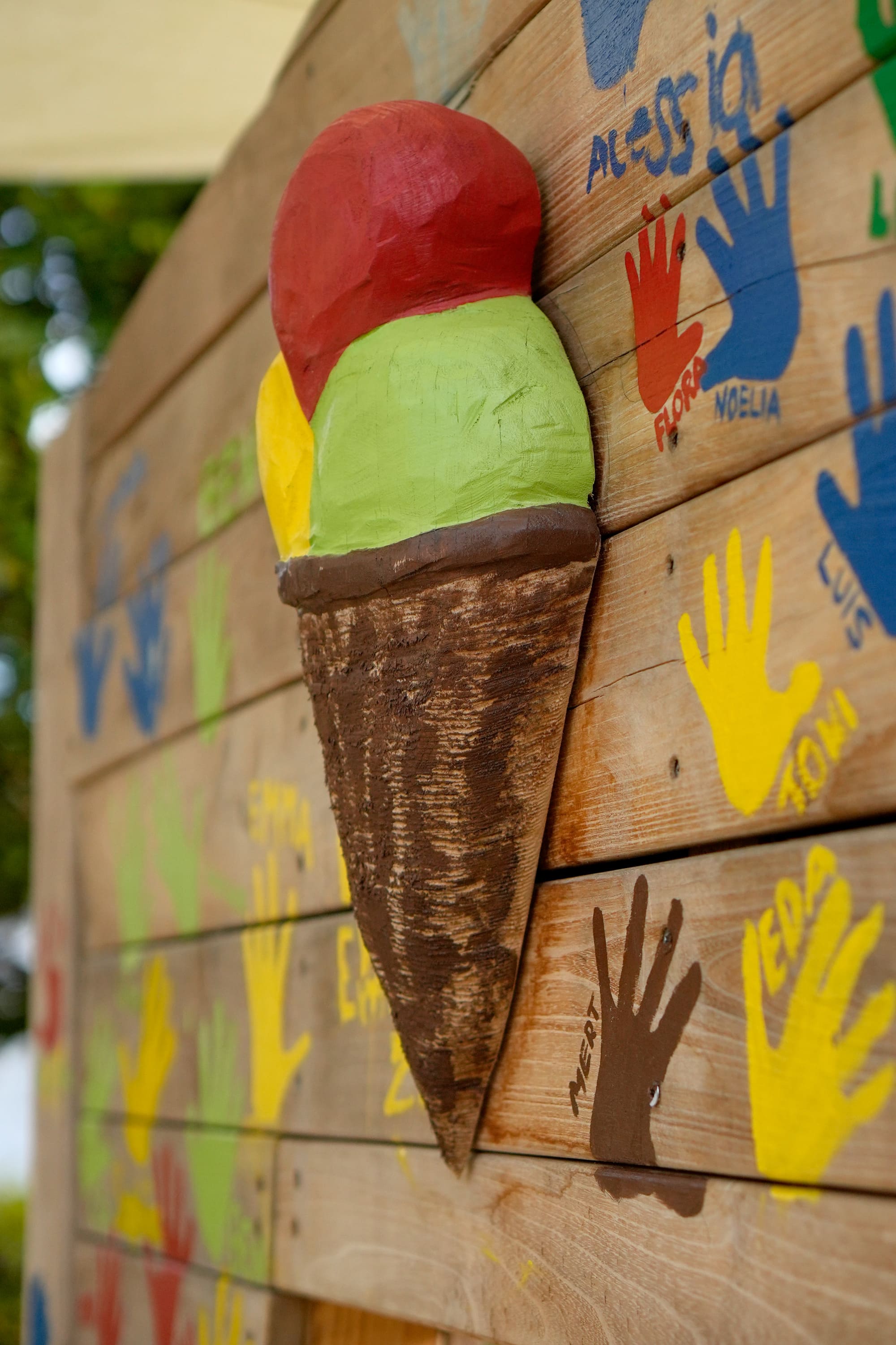 Auf einer mit bunten Handabdrücken bemalten Holzwand ist eine dreidimensionale Eistüte aus Holz mit drei Kugeln in Rot, Grün und Gelb angebracht.