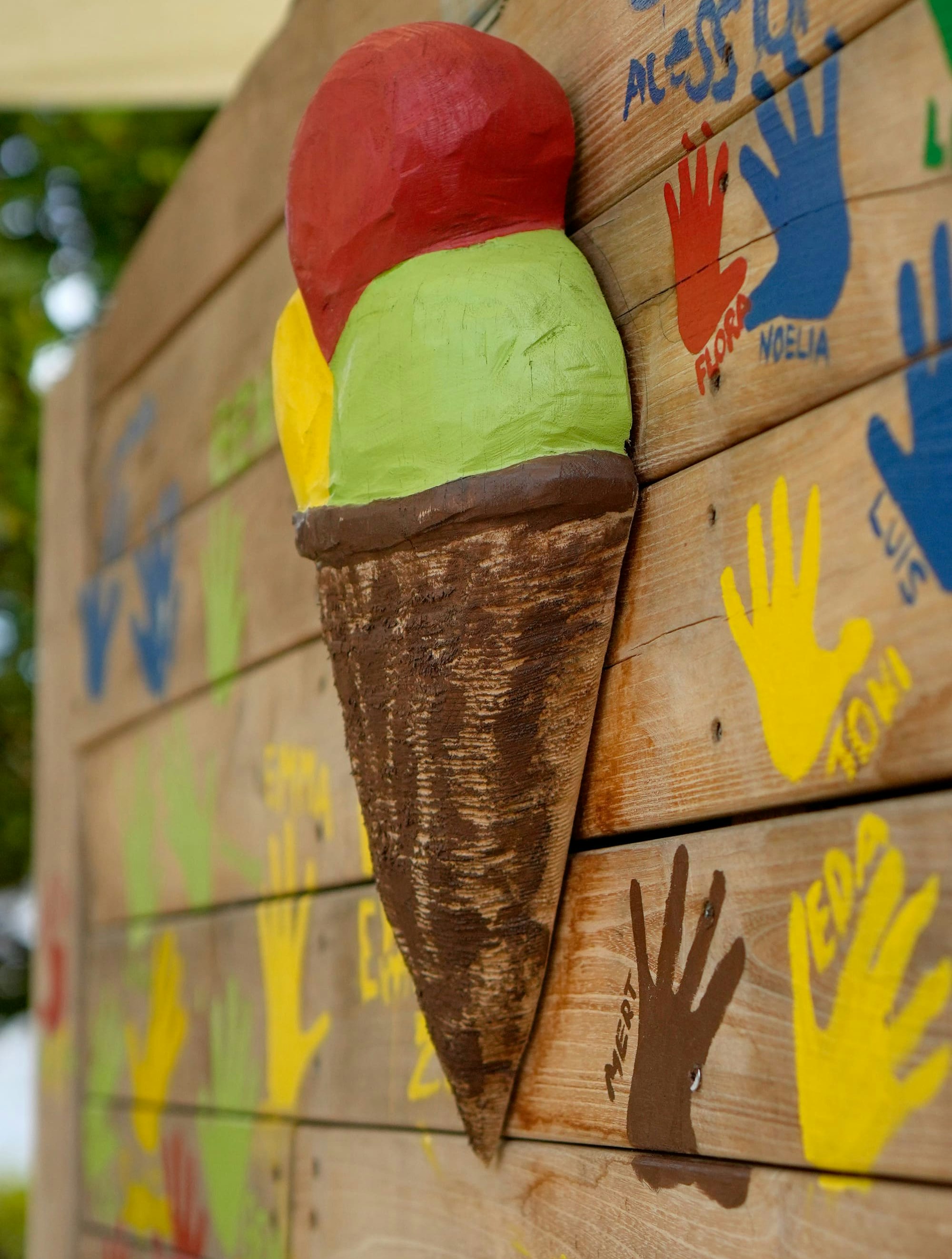 Auf einer mit bunten Handabdrücken bemalten Holzwand ist eine dreidimensionale Eistüte aus Holz mit drei Kugeln in Rot, Grün und Gelb angebracht.