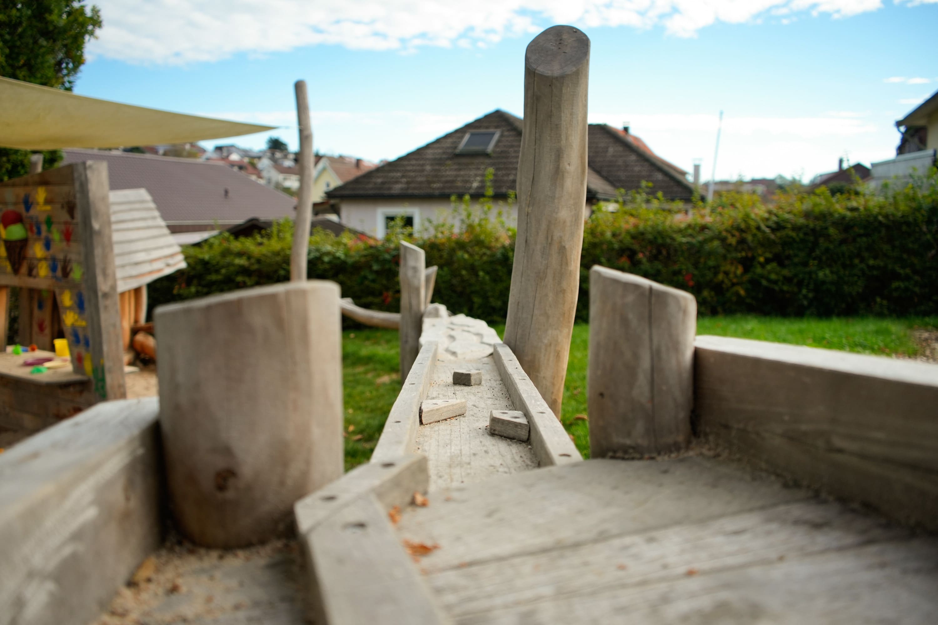 Holzspielgerät mit Schwebebalken und senkrechten Pfosten in einer Hinterhofumgebung. Im Hintergrund sind Häuser und Büsche unter einem teilweise bewölkten Himmel zu sehen.