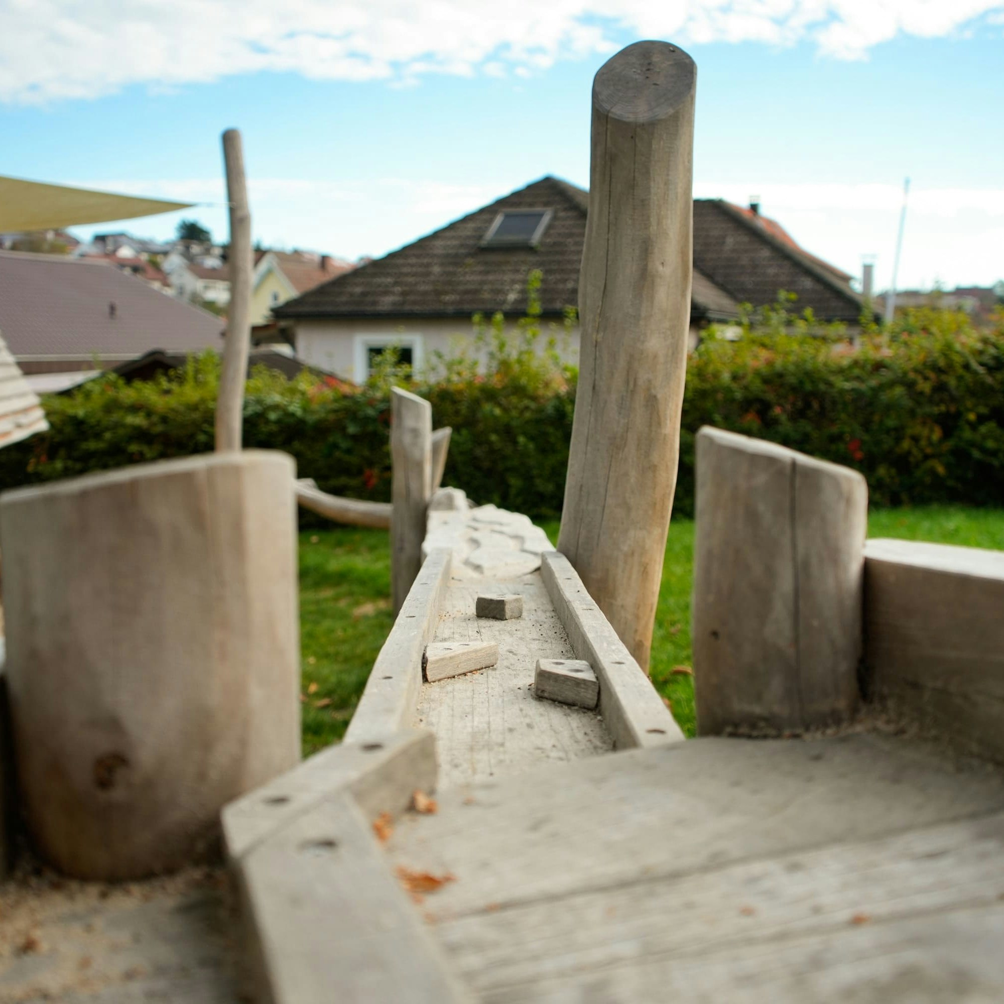 Holzspielgerät mit Schwebebalken und senkrechten Pfosten in einer Hinterhofumgebung. Im Hintergrund sind Häuser und Büsche unter einem teilweise bewölkten Himmel zu sehen.
