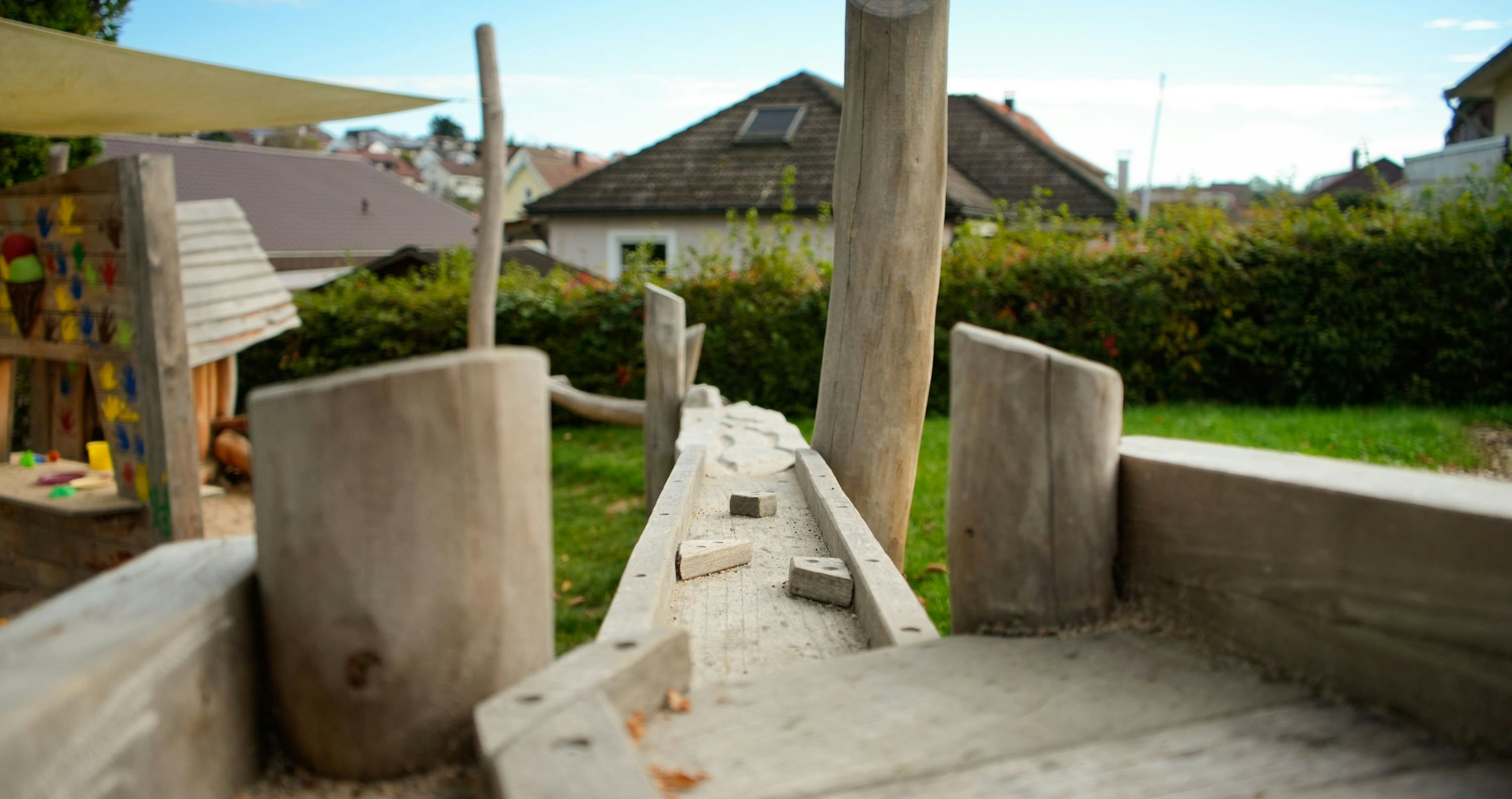 Holzspielgerät mit Schwebebalken und senkrechten Pfosten in einer Hinterhofumgebung. Im Hintergrund sind Häuser und Büsche unter einem teilweise bewölkten Himmel zu sehen.