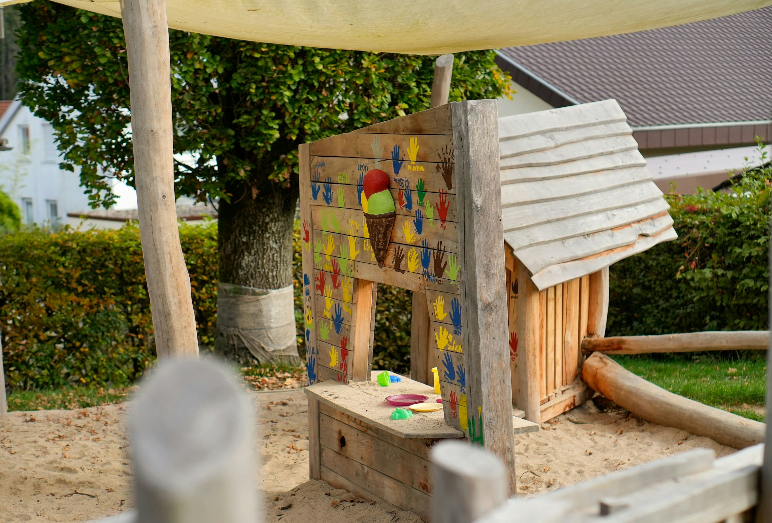 Holzspielstruktur mit einer bunten Tafel mit einer aufgemalten Eistüte und Handabdrücken auf einem Sandspielplatz, umgeben von Bäumen und einer kleinen hausähnlichen Struktur.