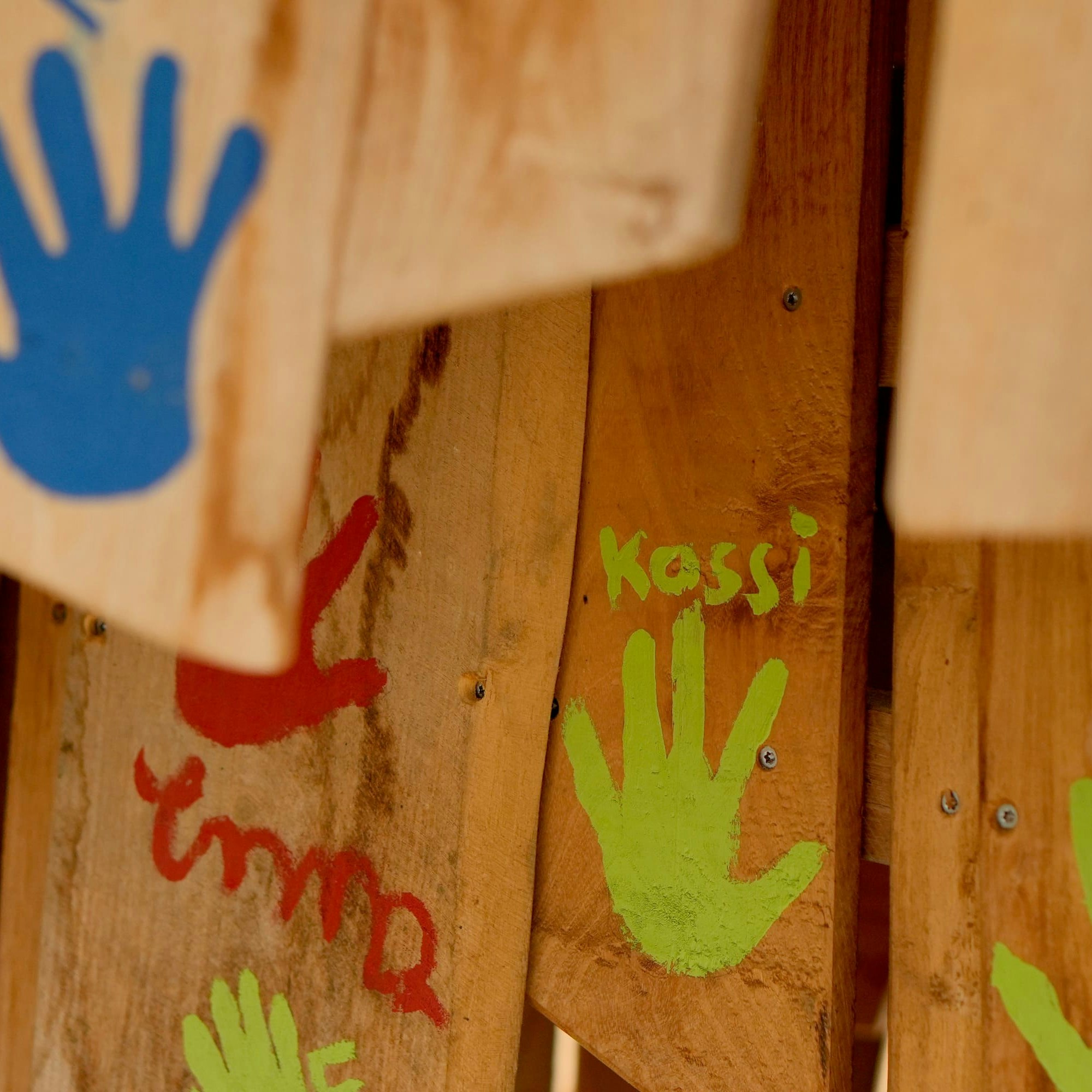 Holzoberfläche mit bunten Handabdrücken: blau, rot, grün und gelb. Der grüne Handabdruck trägt die Aufschrift „Kassi“.