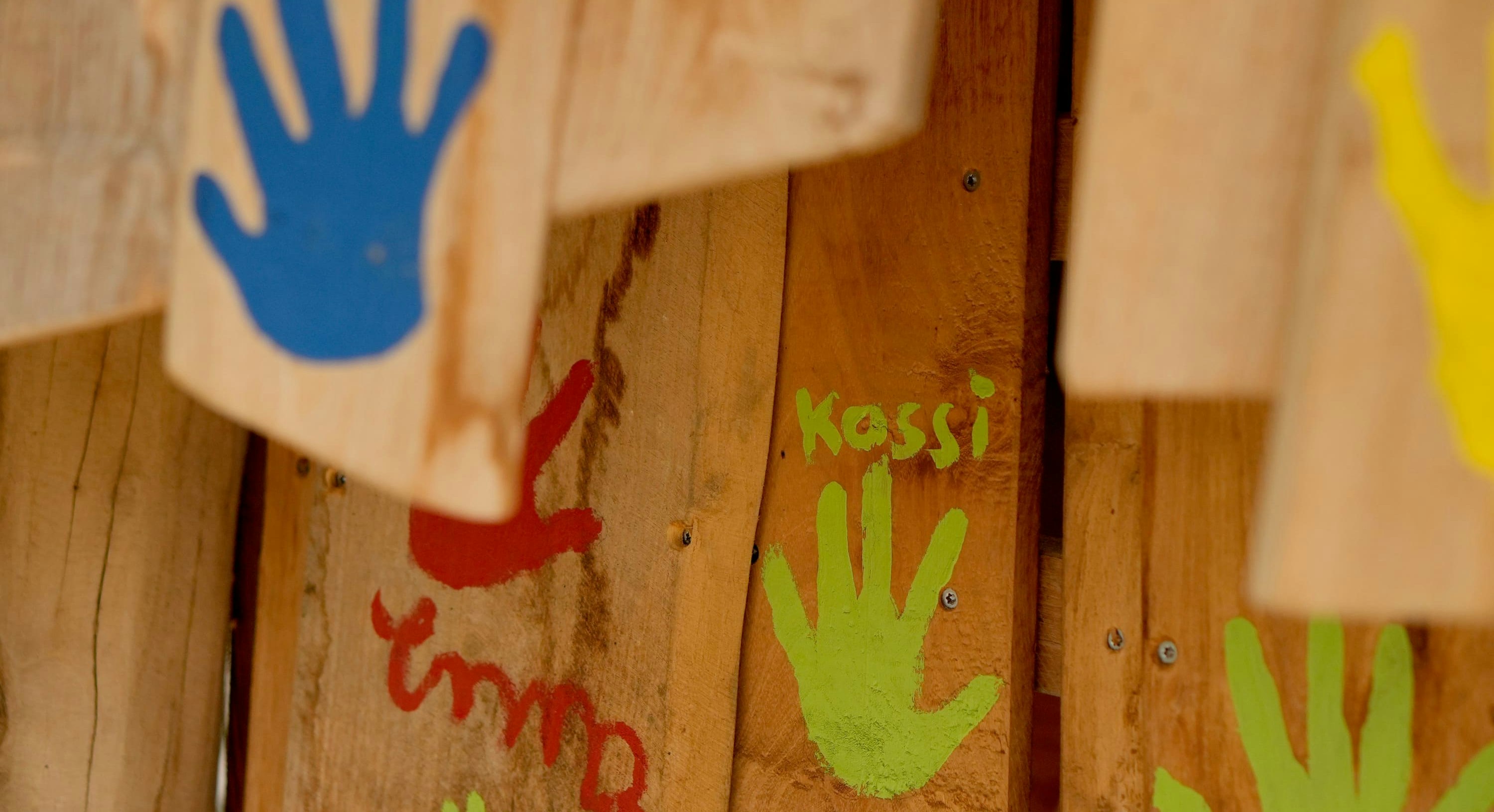 Holzoberfläche mit bunten Handabdrücken: blau, rot, grün und gelb. Der grüne Handabdruck trägt die Aufschrift „Kassi“.