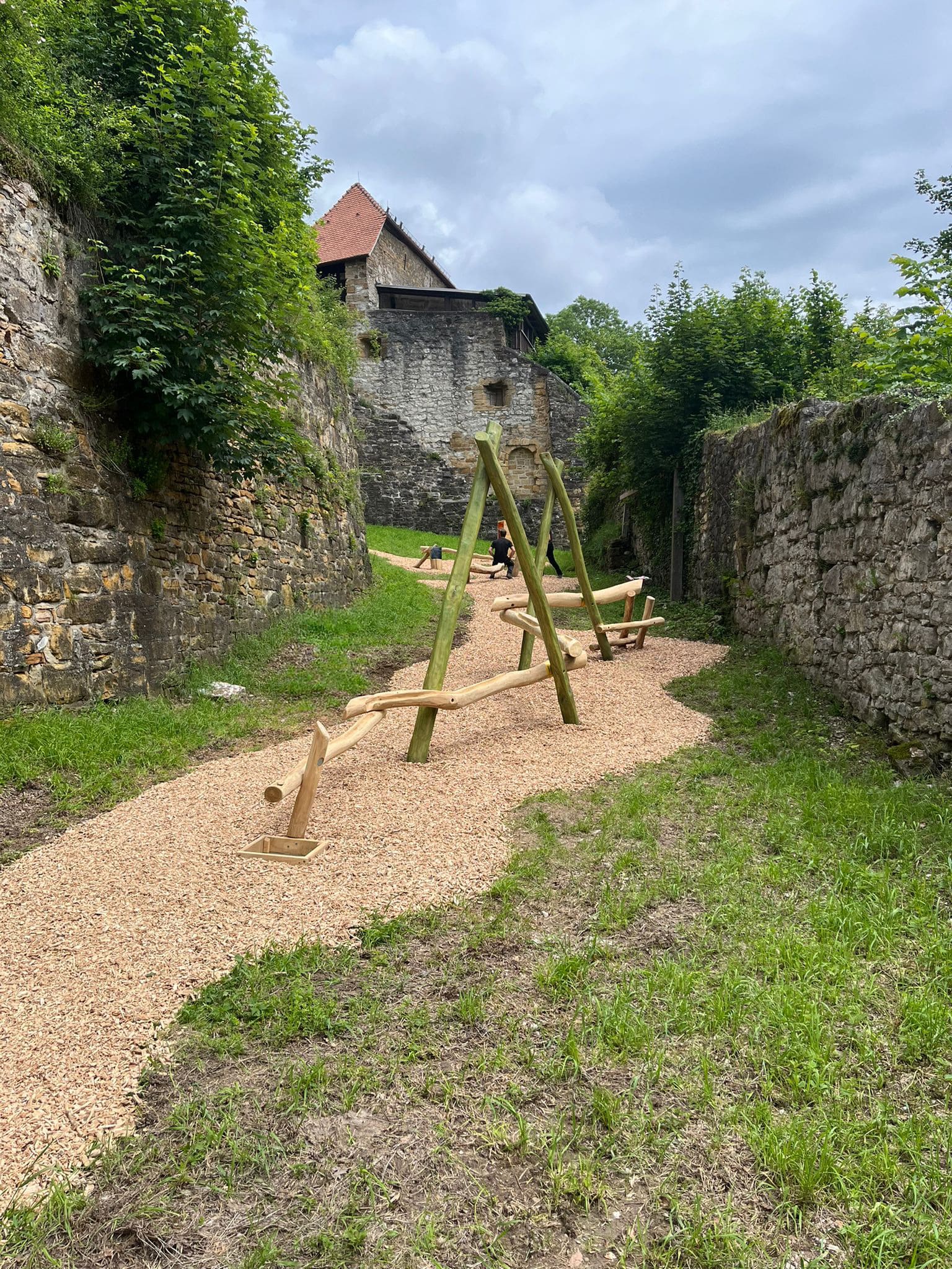 Holzwippen auf einem Kiesweg, umgeben von Steinmauern und Grün, mit einem rustikalen Gebäude im Hintergrund.