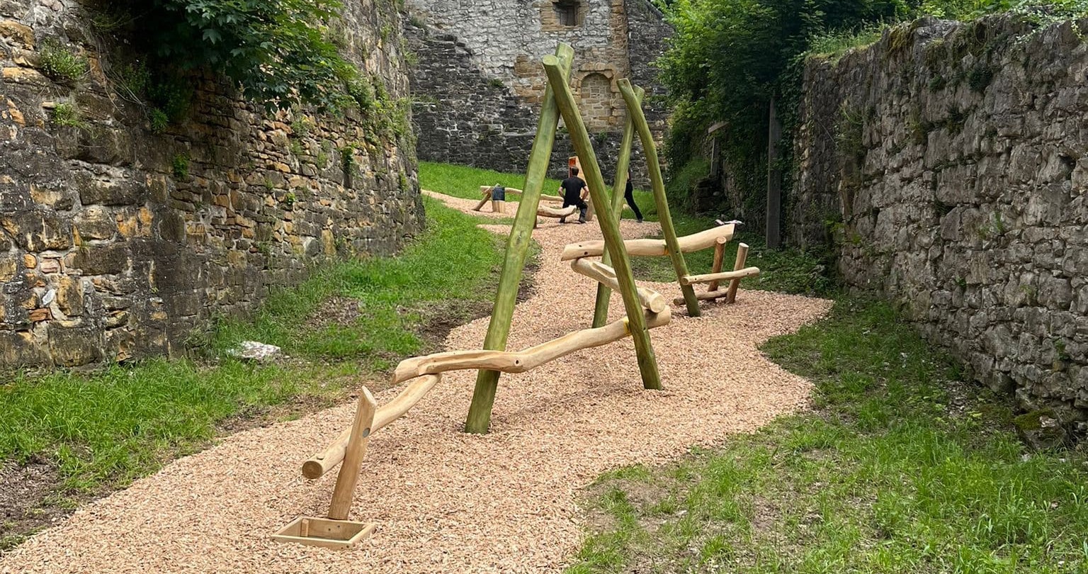 Holzwippen auf einem Kiesweg, umgeben von Steinmauern und Grün, mit einem rustikalen Gebäude im Hintergrund.