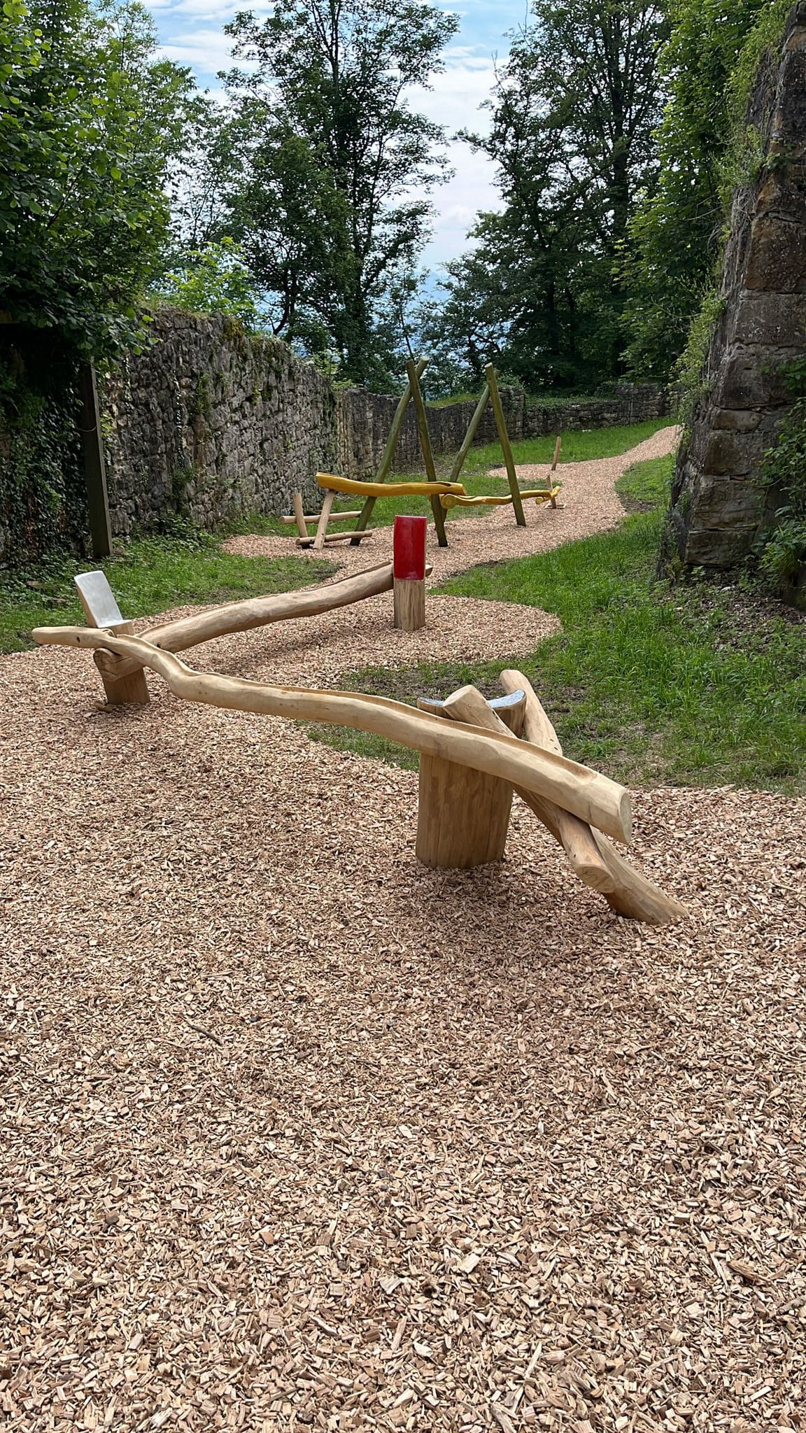 Hölzerne Schwebebalken auf einem Kiesweg, umgeben von Bäumen und Steinmauern.