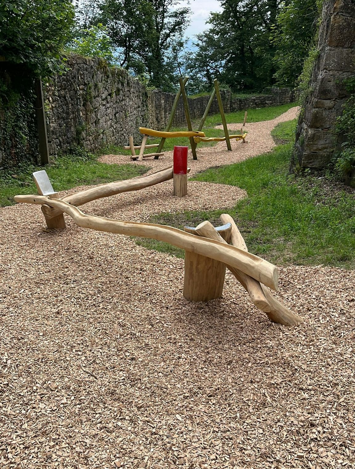 Hölzerne Schwebebalken auf einem Kiesweg, umgeben von Bäumen und Steinmauern.