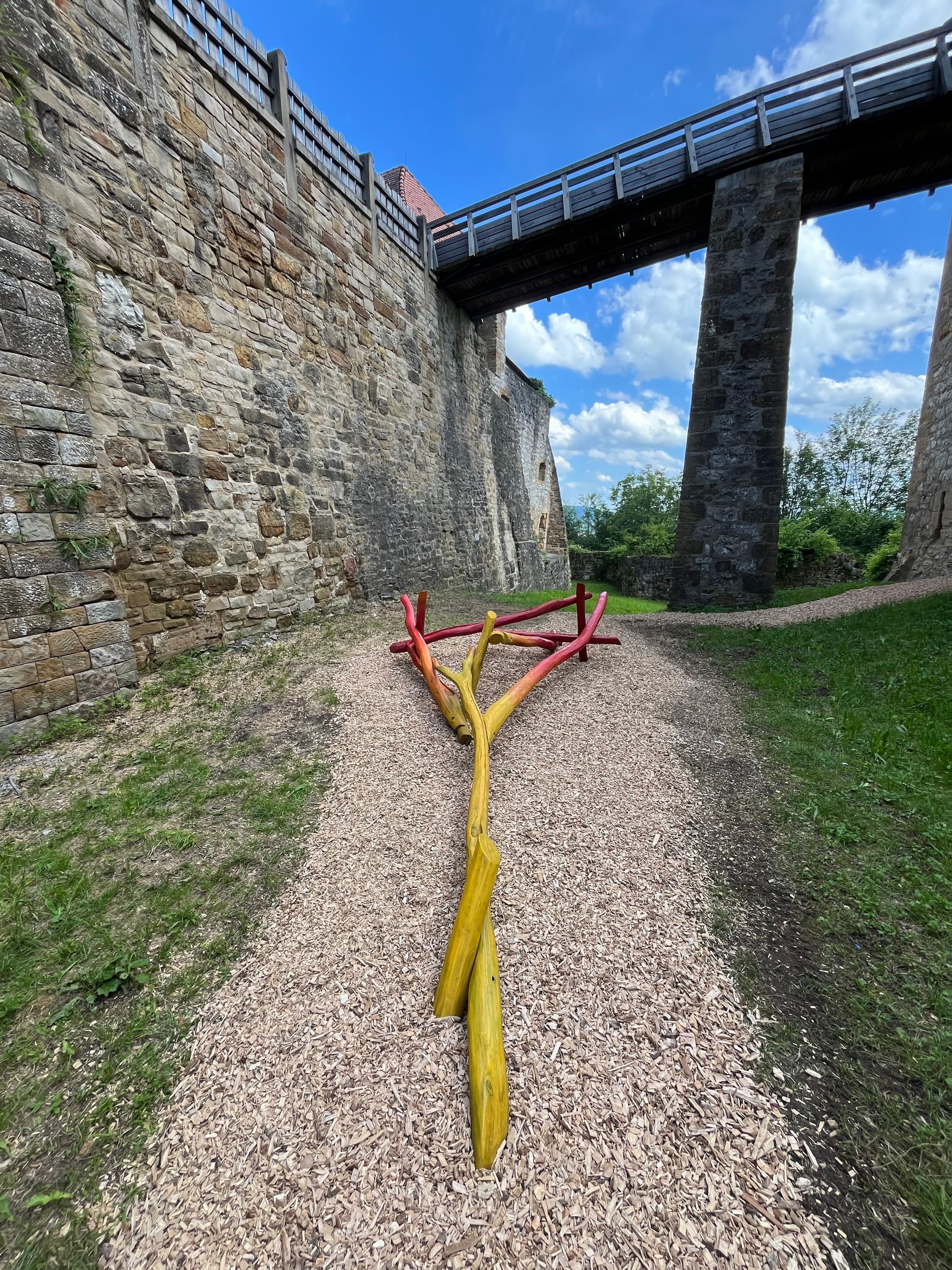 Bunte Holzbalken, angeordnet auf einem Kiesweg unter einer Steinbrücke in der Nähe einer historischen Steinmauer.