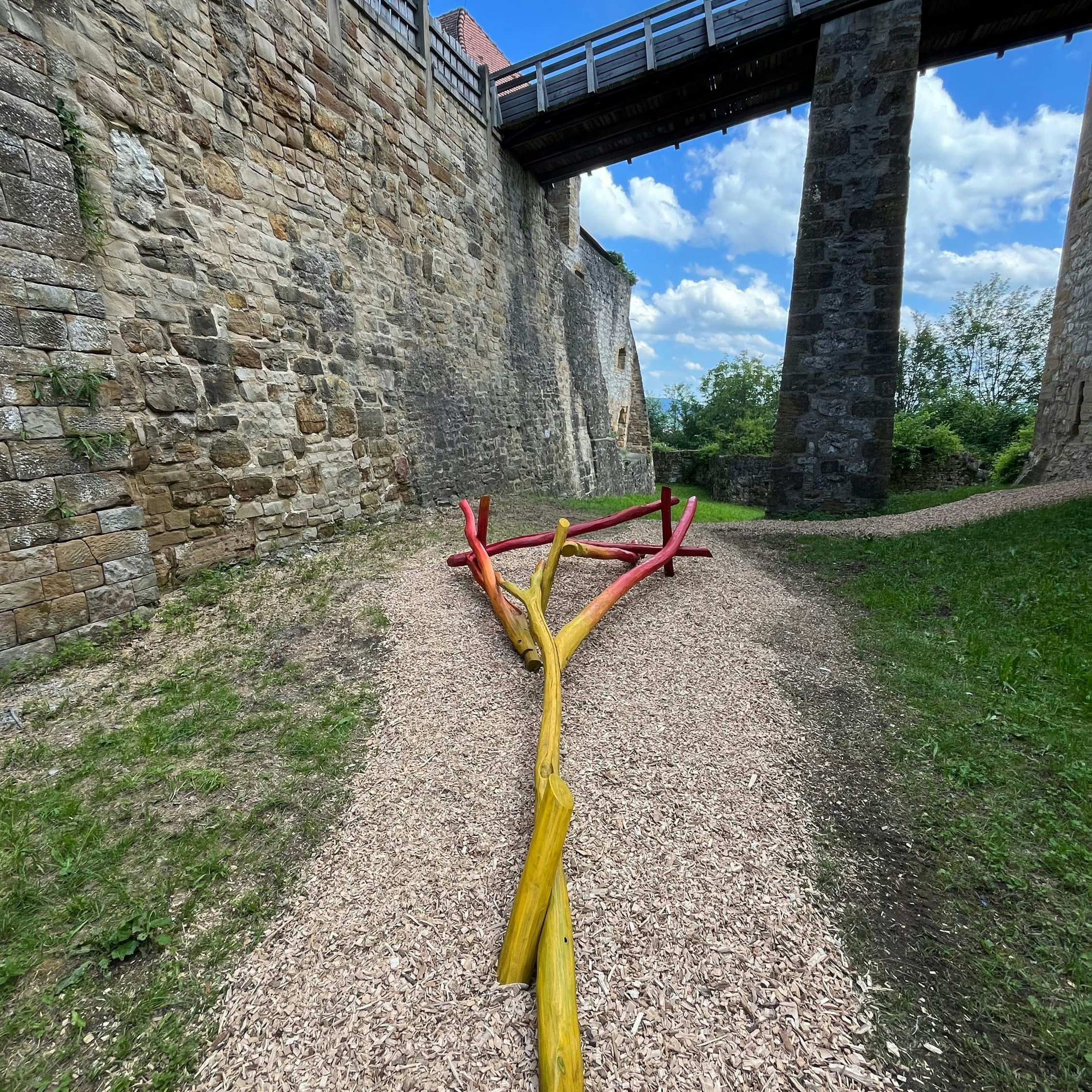 Bunte Holzbalken, angeordnet auf einem Kiesweg unter einer Steinbrücke in der Nähe einer historischen Steinmauer.
