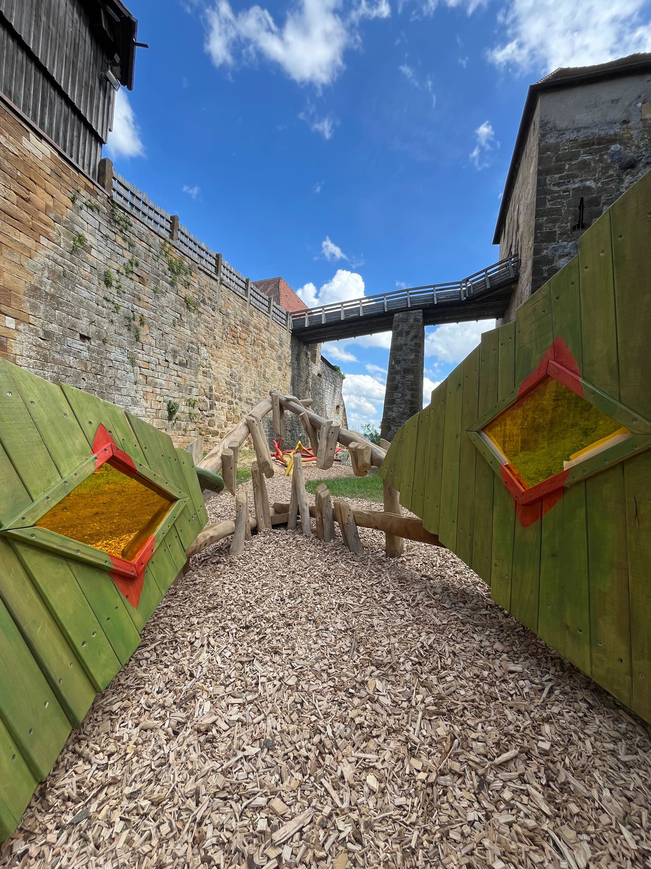 Kinderspielplatz mit Klettergerüst aus Holz und grünen Platten, auf Holzspänen aufgebaut. Blauer Himmel mit Wolken und Steinmauer im Hintergrund.