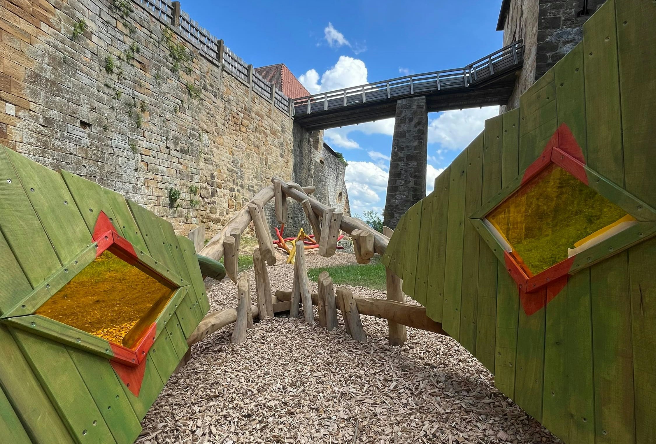 Kinderspielplatz mit Klettergerüst aus Holz und grünen Platten, auf Holzspänen aufgebaut. Blauer Himmel mit Wolken und Steinmauer im Hintergrund.