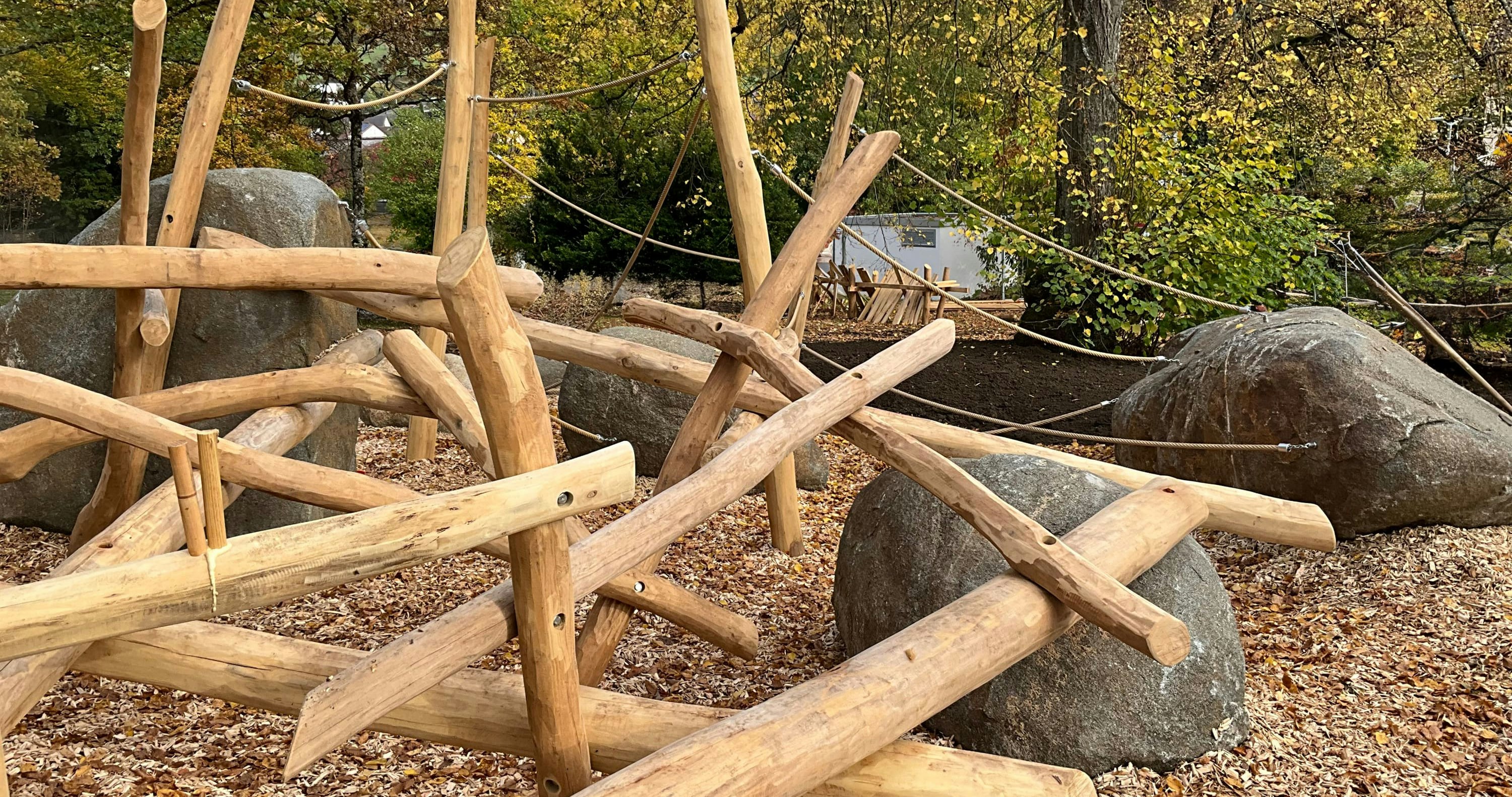 Hölzernes Klettergerüst mit Seilen und großen Felsbrocken in einer Parklandschaft, umgeben von Herbstbäumen und gefallenen Blättern.