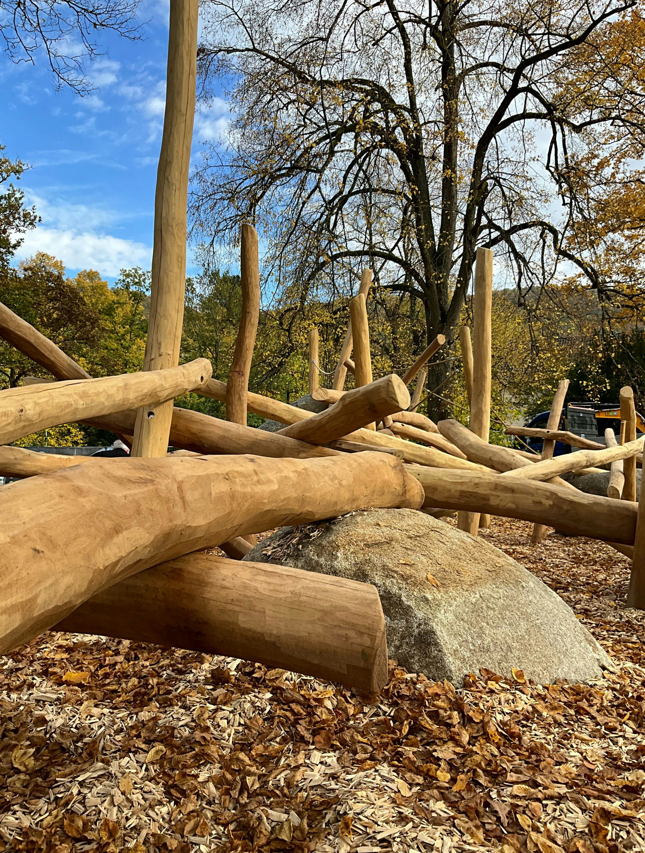 Holzspielstruktur mit kreuz und quer angeordneten Baumstämmen auf einem mit Blättern bedeckten Waldboden. Im Hintergrund sind Bäume mit Herbstlaub unter einem blauen Himmel zu sehen.