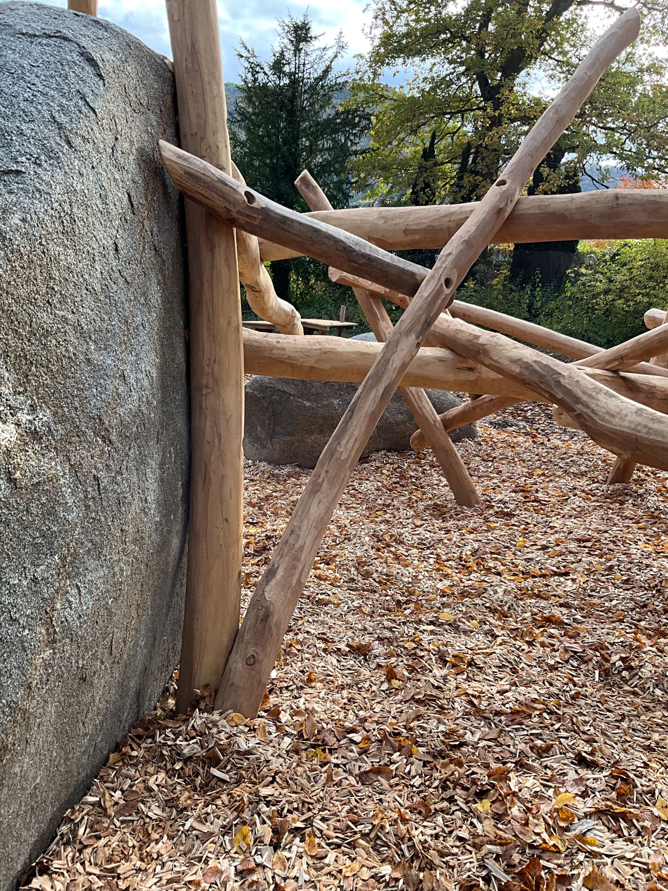 Außenstruktur mit Holzbalken und Steinmauer, auf einem mit Holzspänen bedeckten und von Bäumen umgebenen Boden.