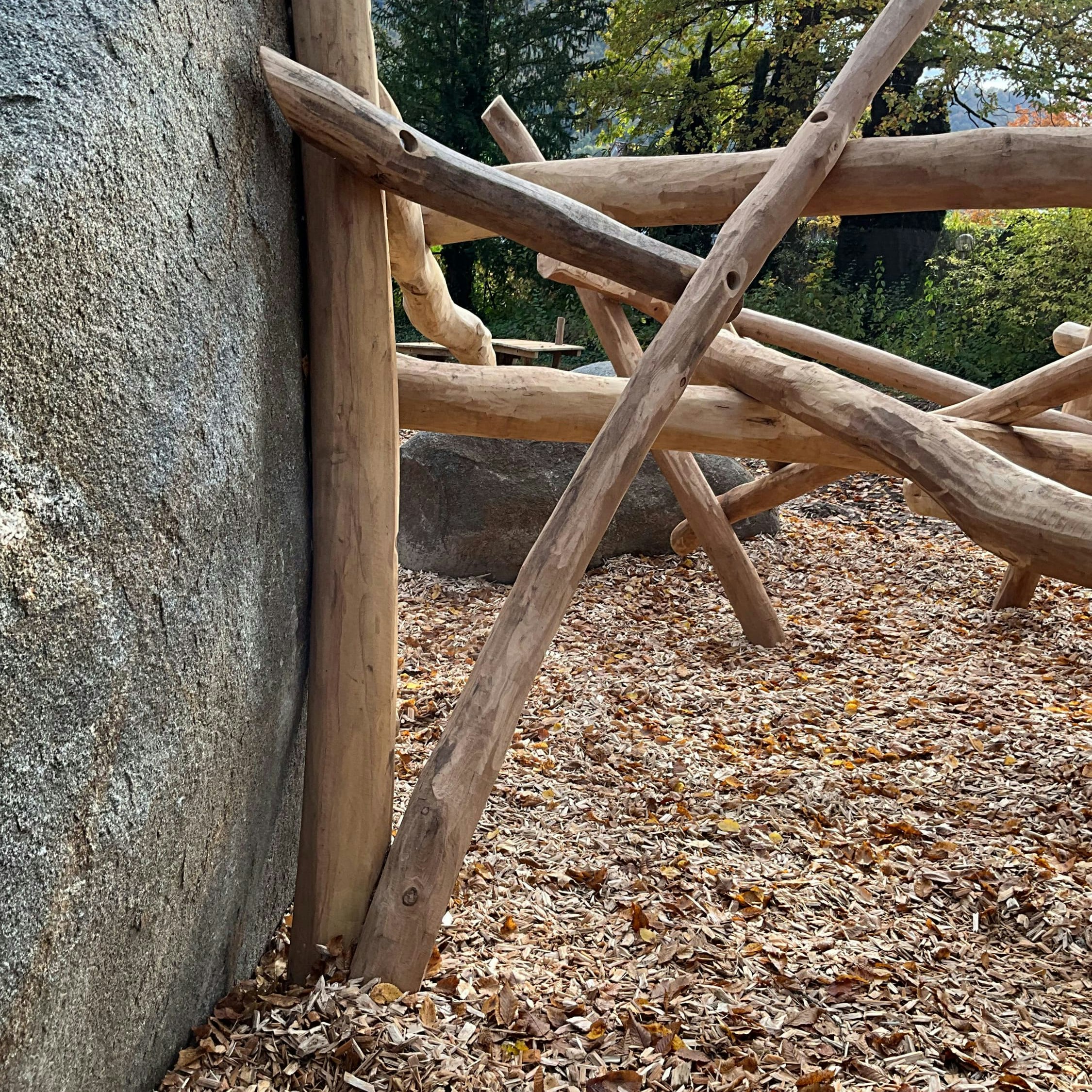 Außenstruktur mit Holzbalken und Steinmauer, auf einem mit Holzspänen bedeckten und von Bäumen umgebenen Boden.