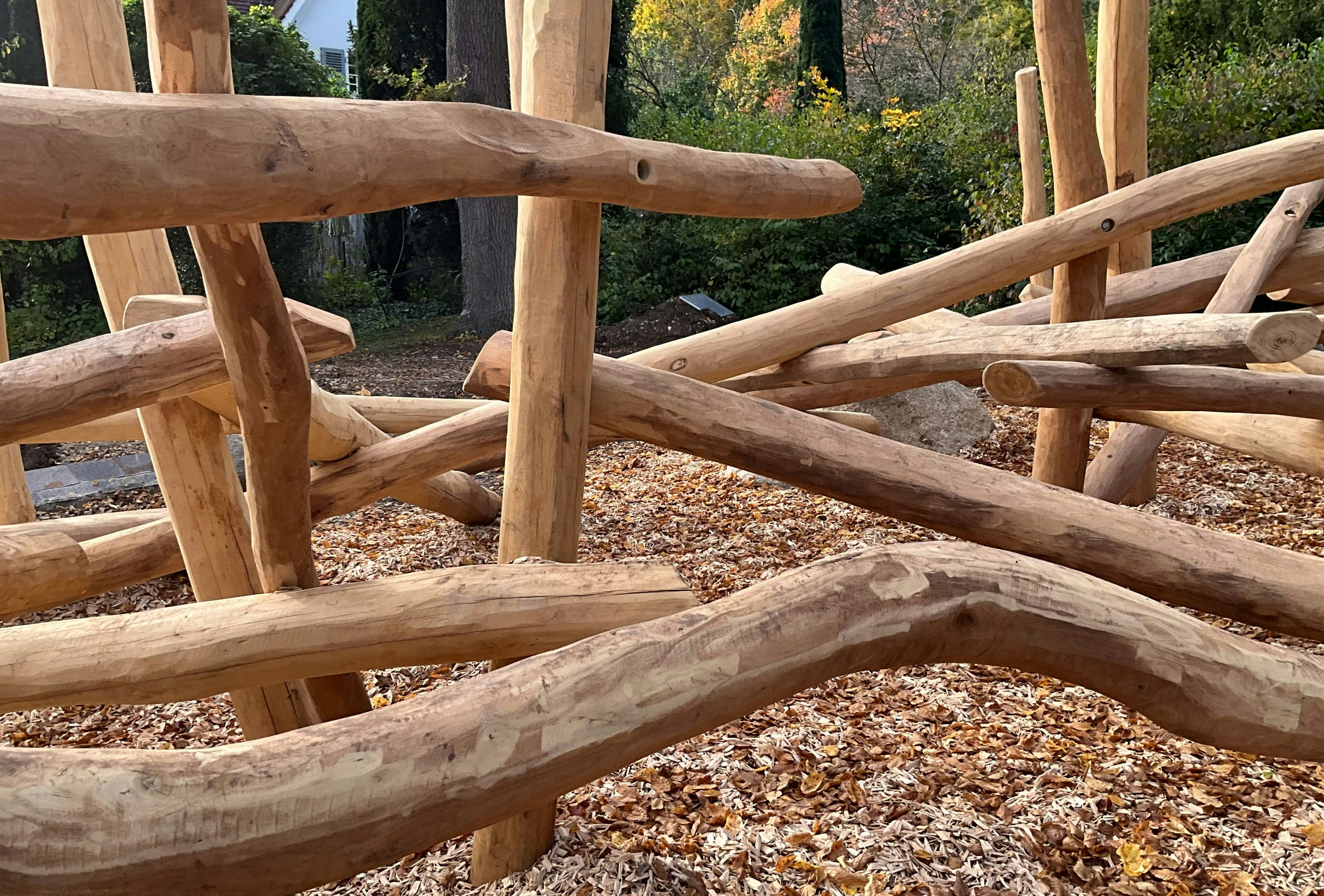 Natürliche Holzkonstruktion mit überlappenden Baumstämmen, umgeben von herbstlichen Blättern, in einem bewaldeten Außenbereich.