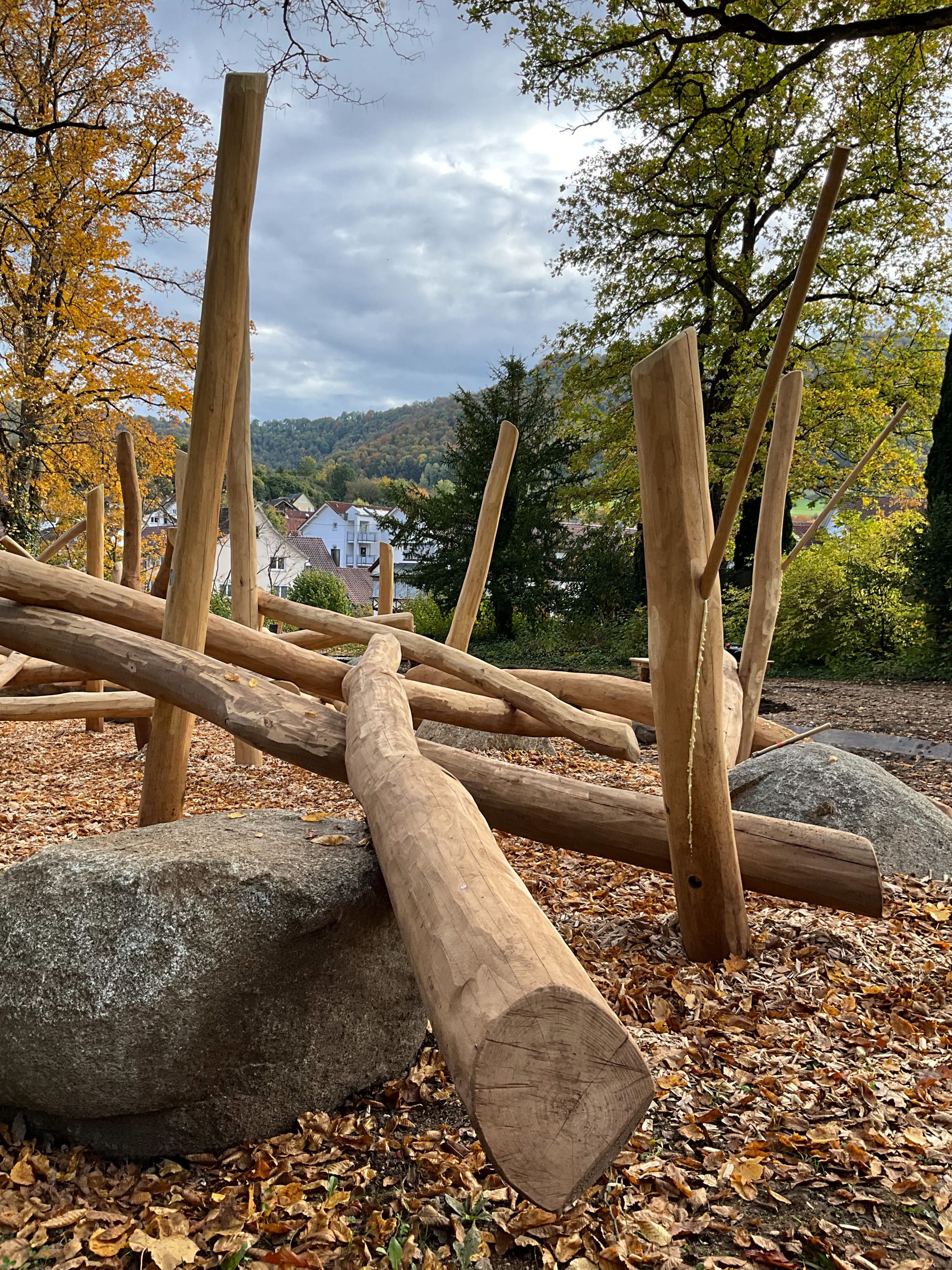 Hölzernes Klettergerüst in einem Park, umgeben von Herbstlaub und Bäumen, mit einem Fernblick auf Häuser und Hügel unter einem bewölkten Himmel.