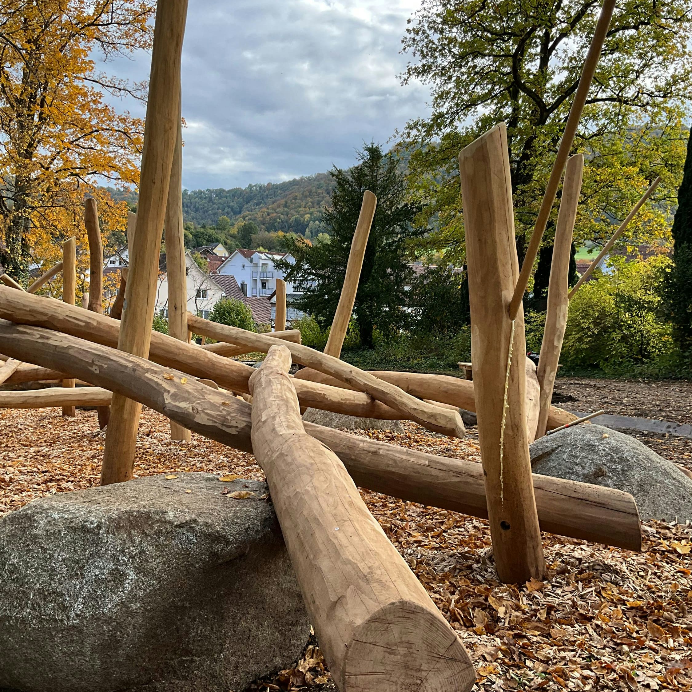 Hölzernes Klettergerüst in einem Park, umgeben von Herbstlaub und Bäumen, mit einem Fernblick auf Häuser und Hügel unter einem bewölkten Himmel.