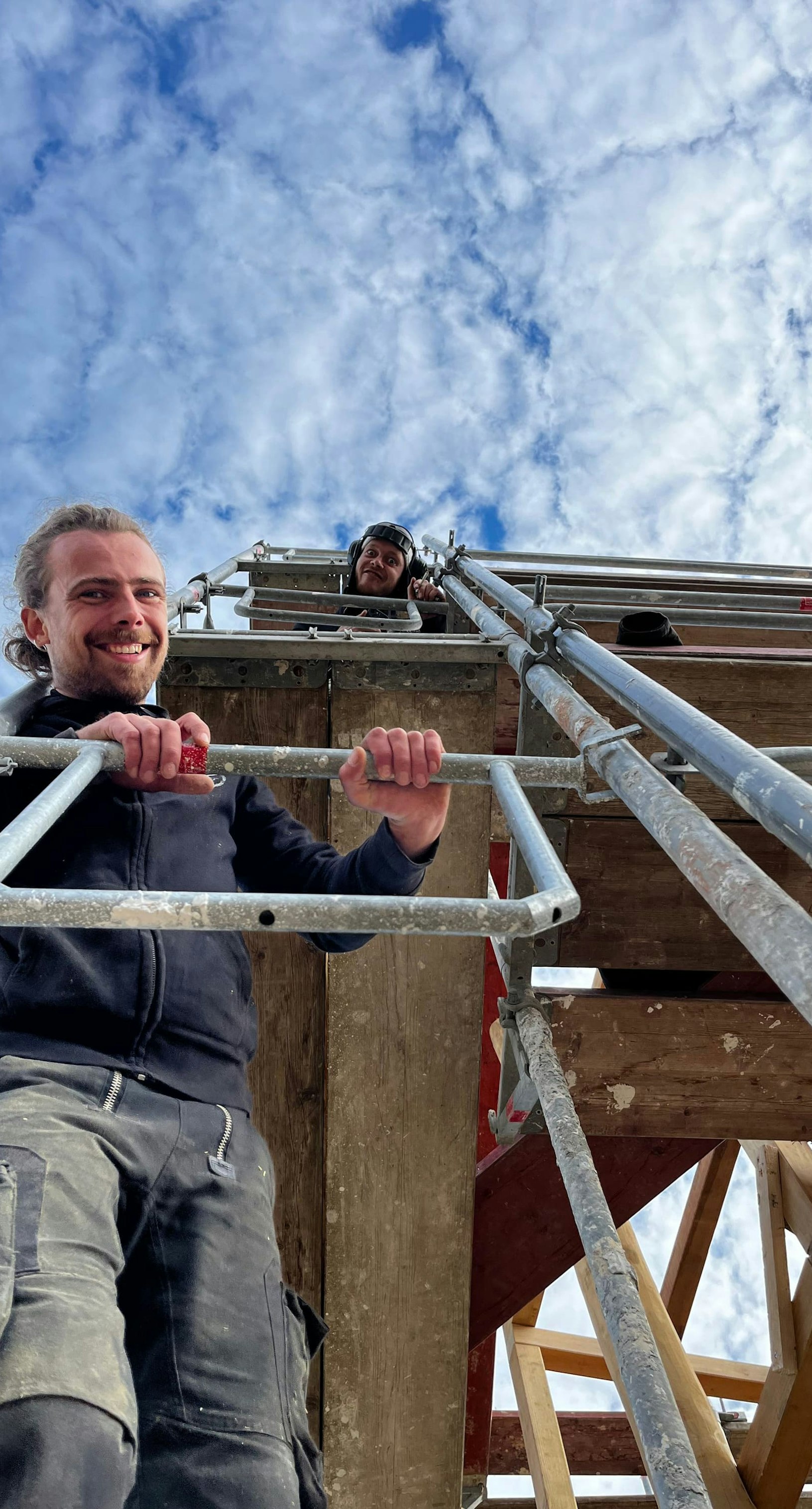 Zwei Arbeiter auf einem Gerüst. Einer lächelt von einer unteren Ebene in die Kamera, der andere steht weiter oben vor der Kulisse eines bewölkten Himmels.