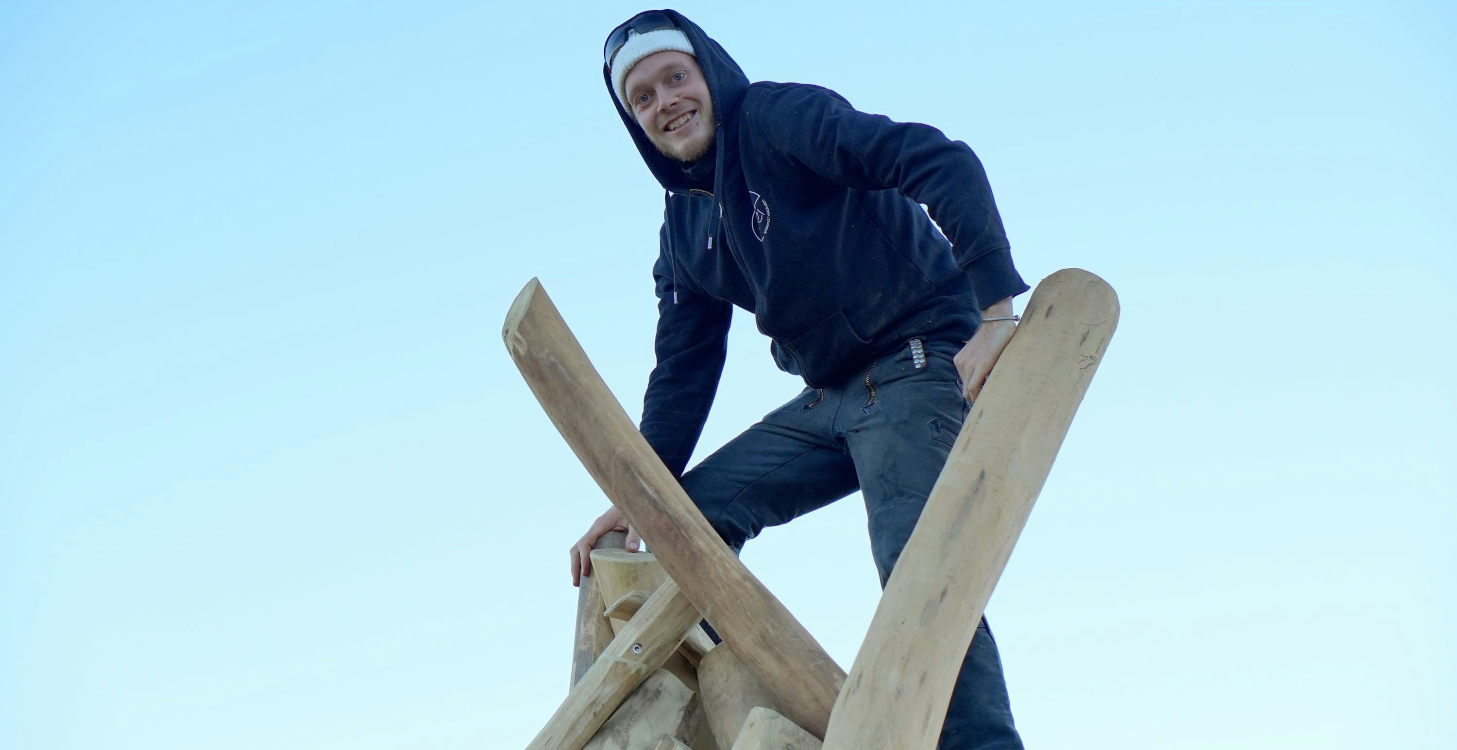 Person, die einen schwarzen Kapuzenpullover und Jeans trägt und vor einem klaren blauen Himmel auf einer Holzkonstruktion steht.
