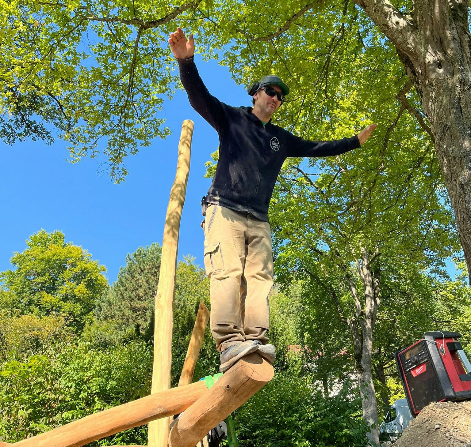 Person balanciert im Freien auf einer Holzkonstruktion, die Arme ausgestreckt, trägt einen schwarzen Pullover, beige Hosen und eine Sonnenbrille. Im Hintergrund Bäume und blauer Himmel.