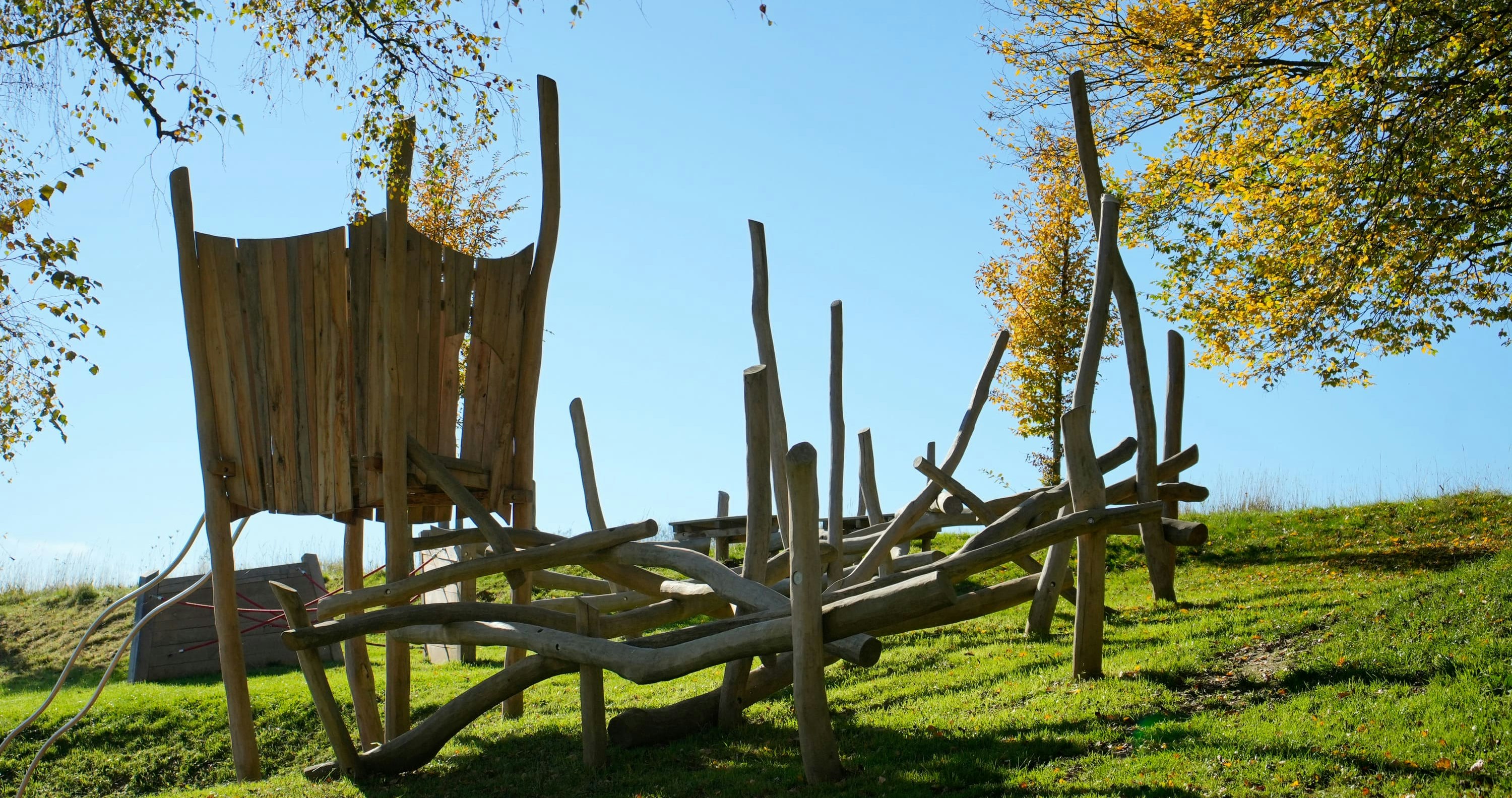 Hölzerne Spielplatzstruktur mit abstraktem Design auf Gras, umgeben von Herbstbäumen unter einem klaren blauen Himmel.