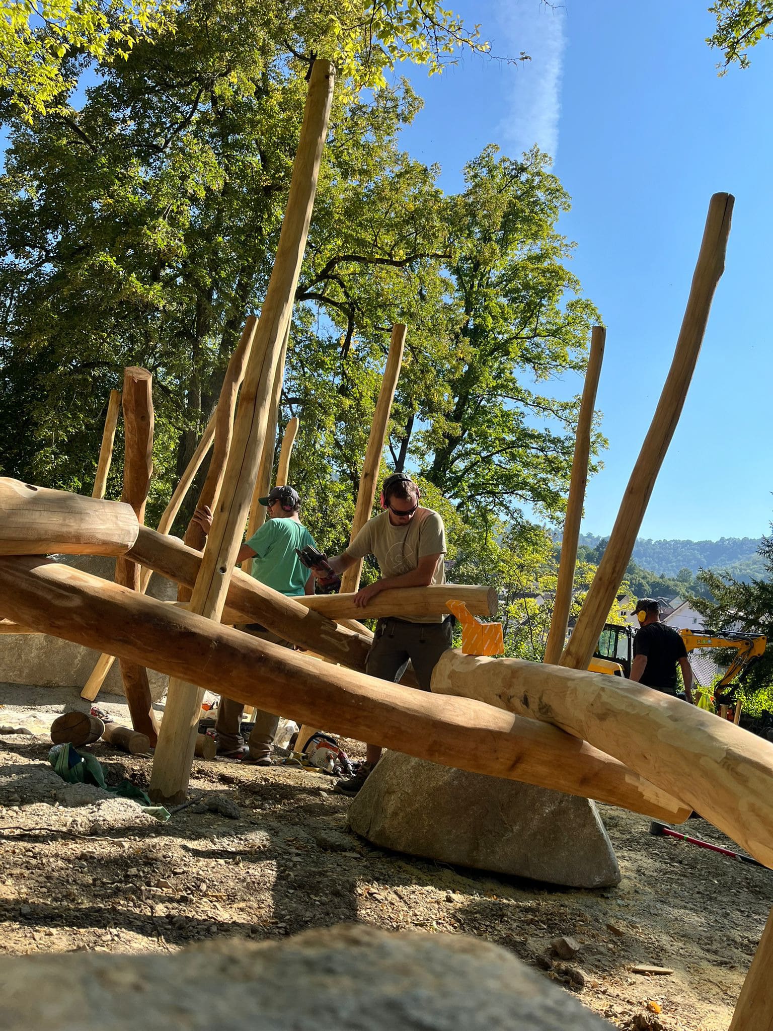 Zwei Personen errichten im Freien, umgeben von Bäumen, eine Holzkonstruktion. Sie arbeiten unter klarem Himmel mit Baumstämmen und Balken.